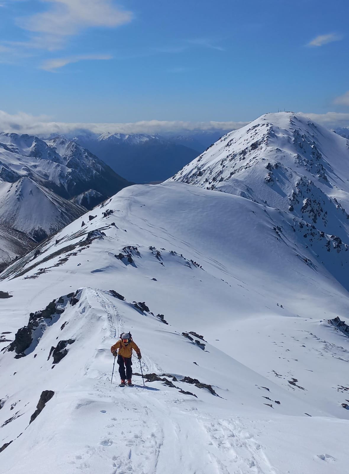 A person hiking in snowy mountain terrain with snow-covered peaks and a blue sky in the background.