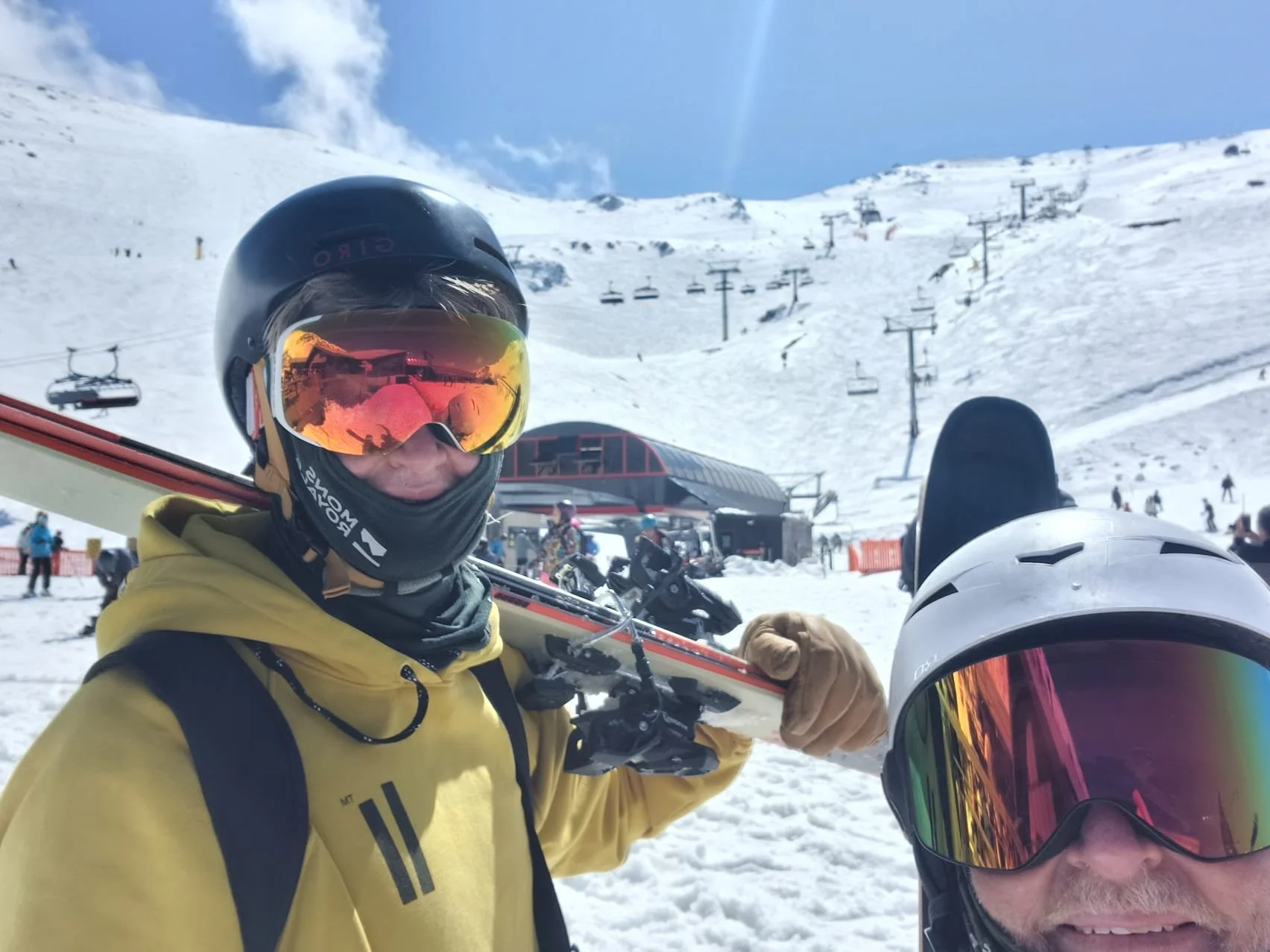 Two people at a snowy ski resort wearing helmets and goggles, with ski lifts and other skiers in the background.