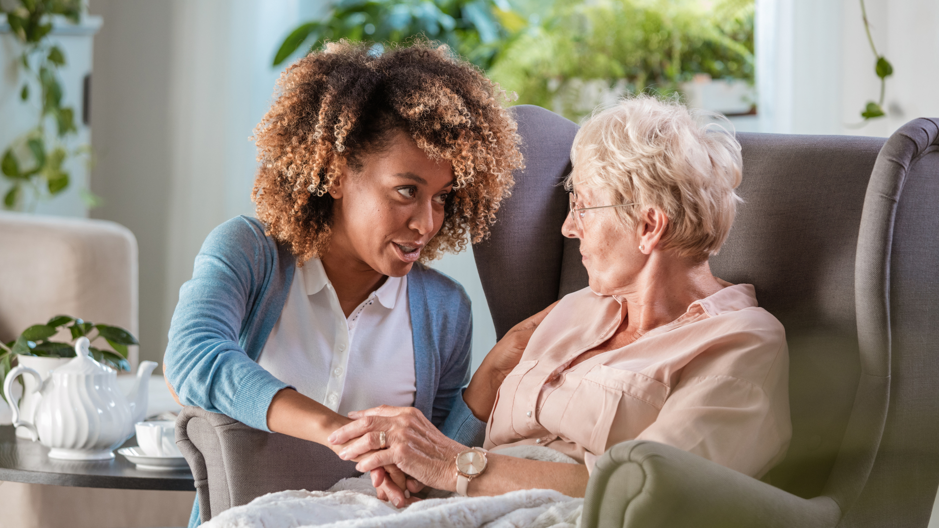 A caregiver talking to an elderly woman seated in a recliner, holding her hand and engaging in conversation.