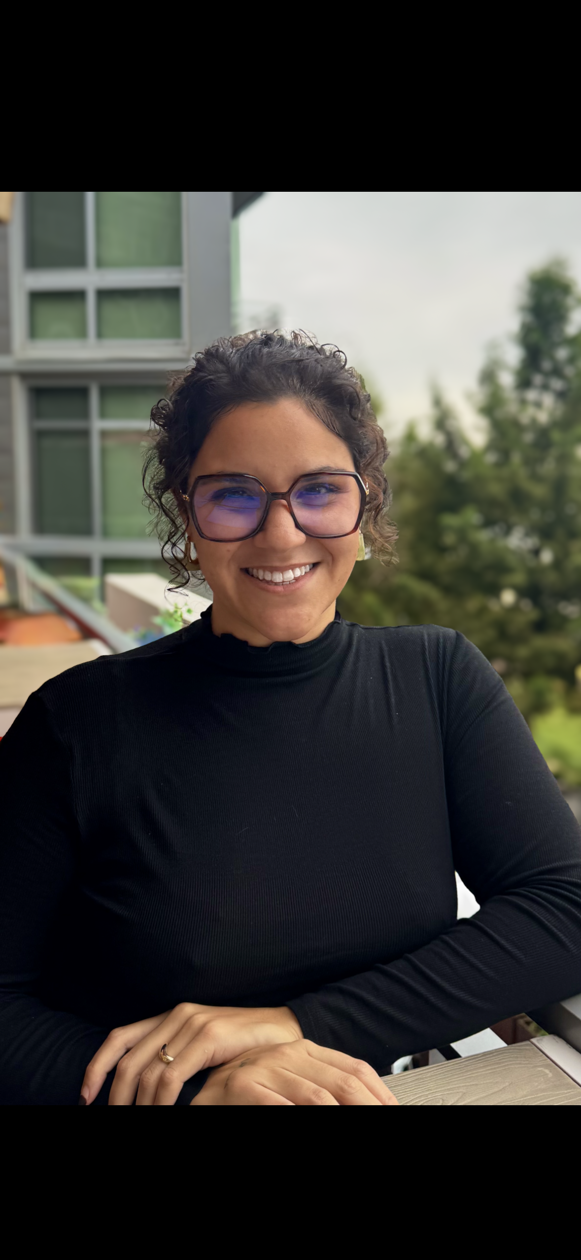 A woman with curly dark hair and glasses smiling at the camera, sitting outdoors with a modern building and trees in the background.