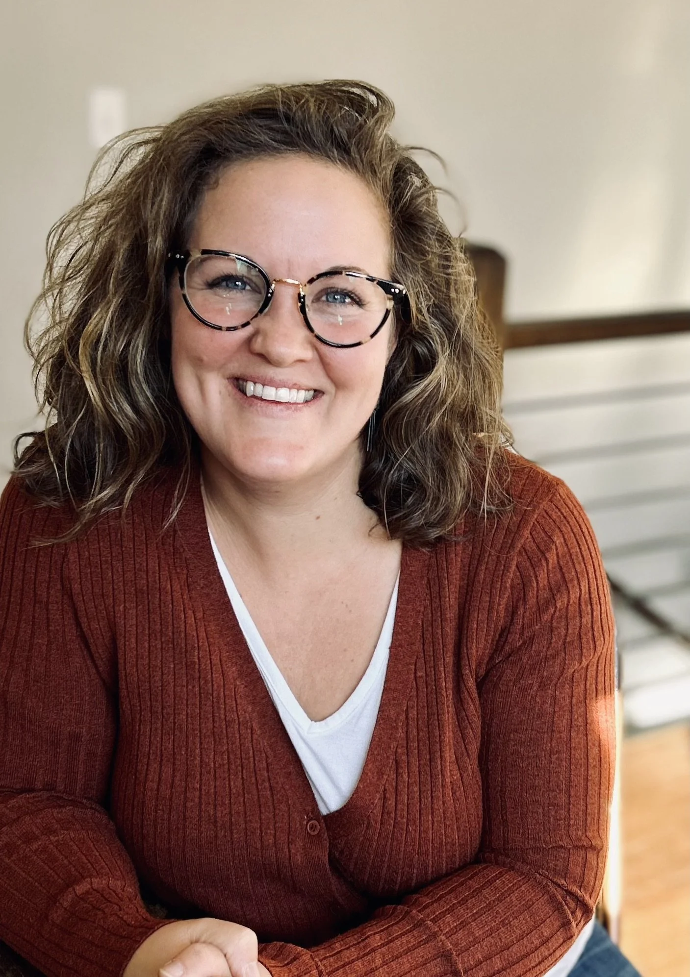 A woman with curly brown hair, glasses, and a white top under a red cardigan, smiling at the camera.