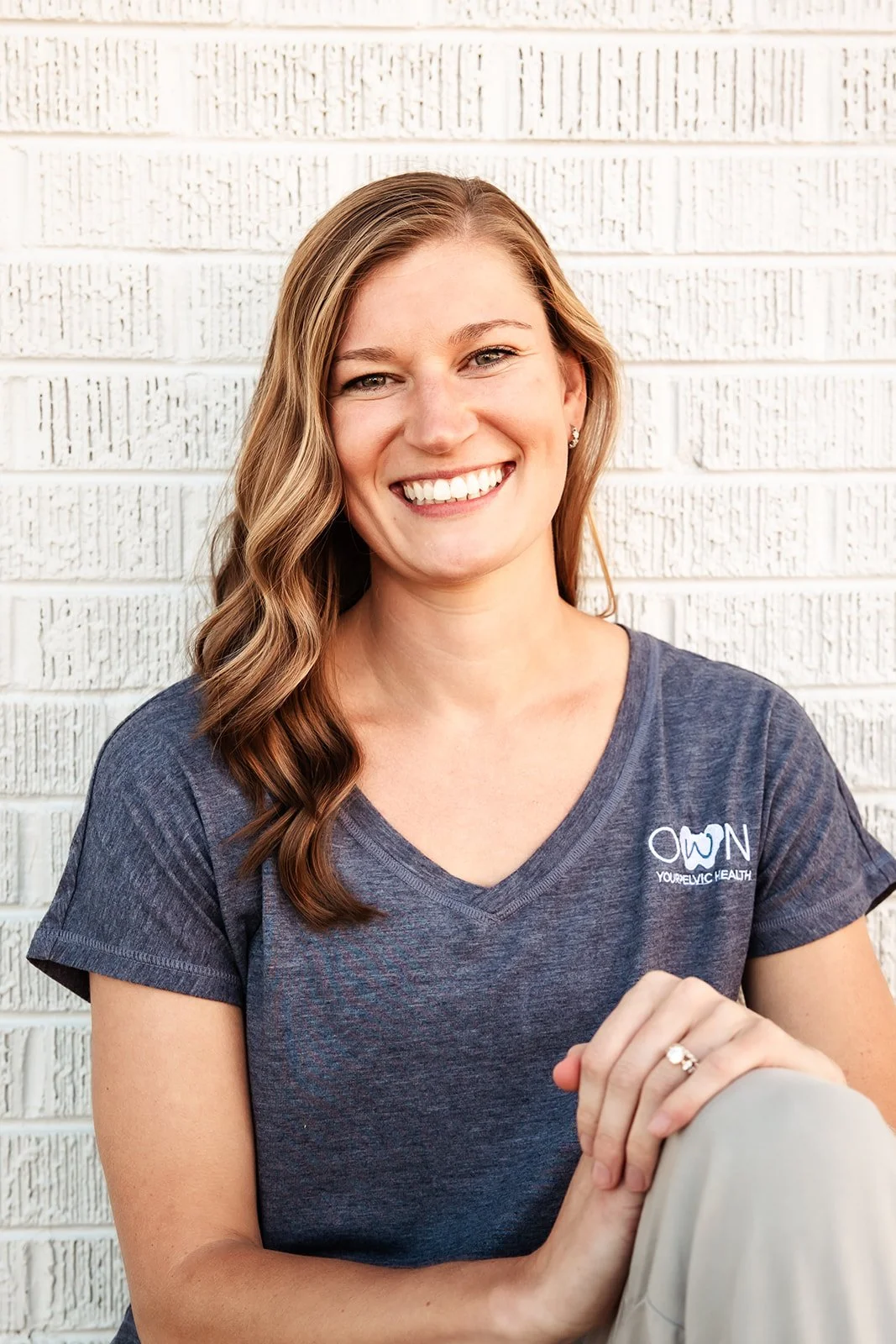 A smiling woman with wavy brown hair wearing a dark gray T-shirt with white text standing against a white brick wall.