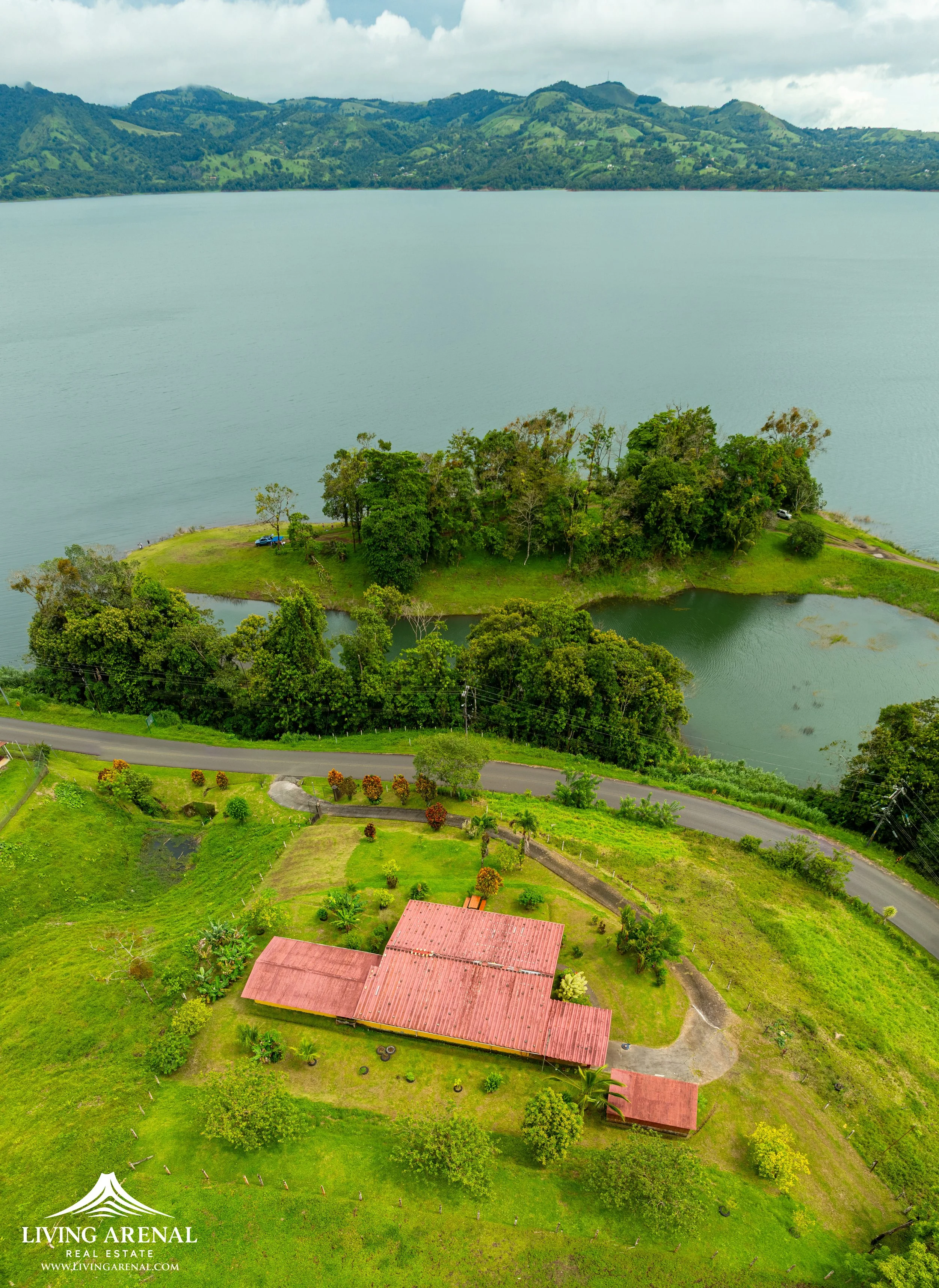 Aerial view of a property with a red-roofed house, surrounded by green lawns and trees, near a body of water with a small island covered in trees, and hills in the background.