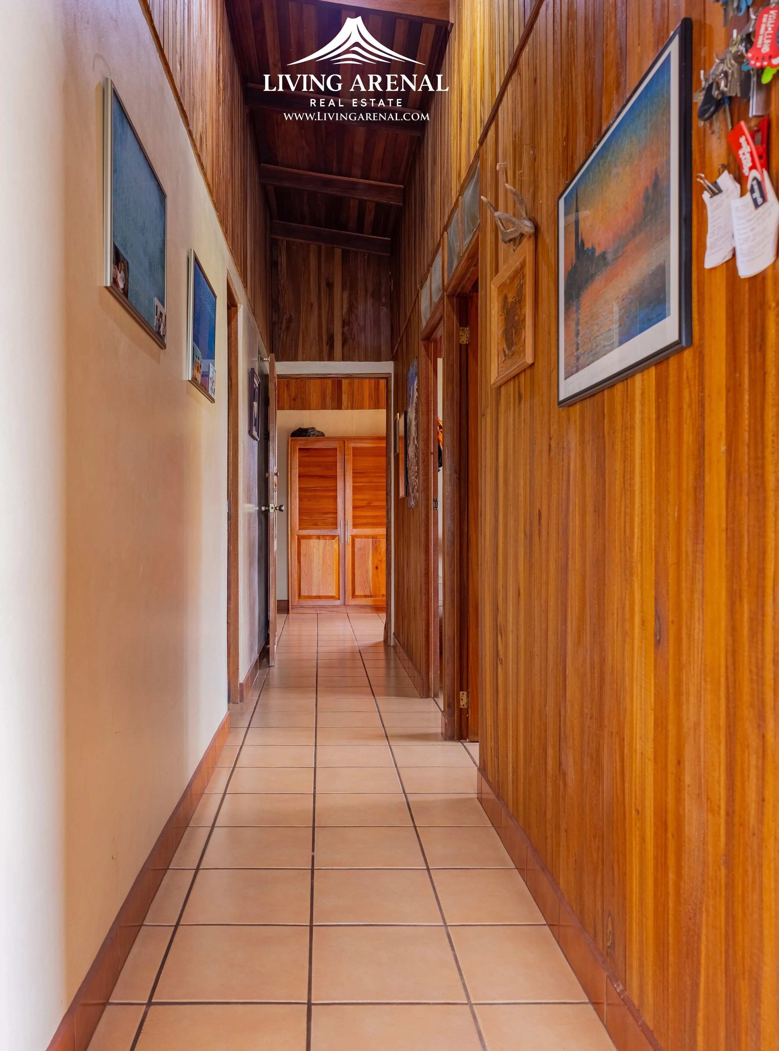 A wooden-paneled hallway with tiled flooring, decorated with framed artwork and a bulletin board, leading to a cabinet at the end.