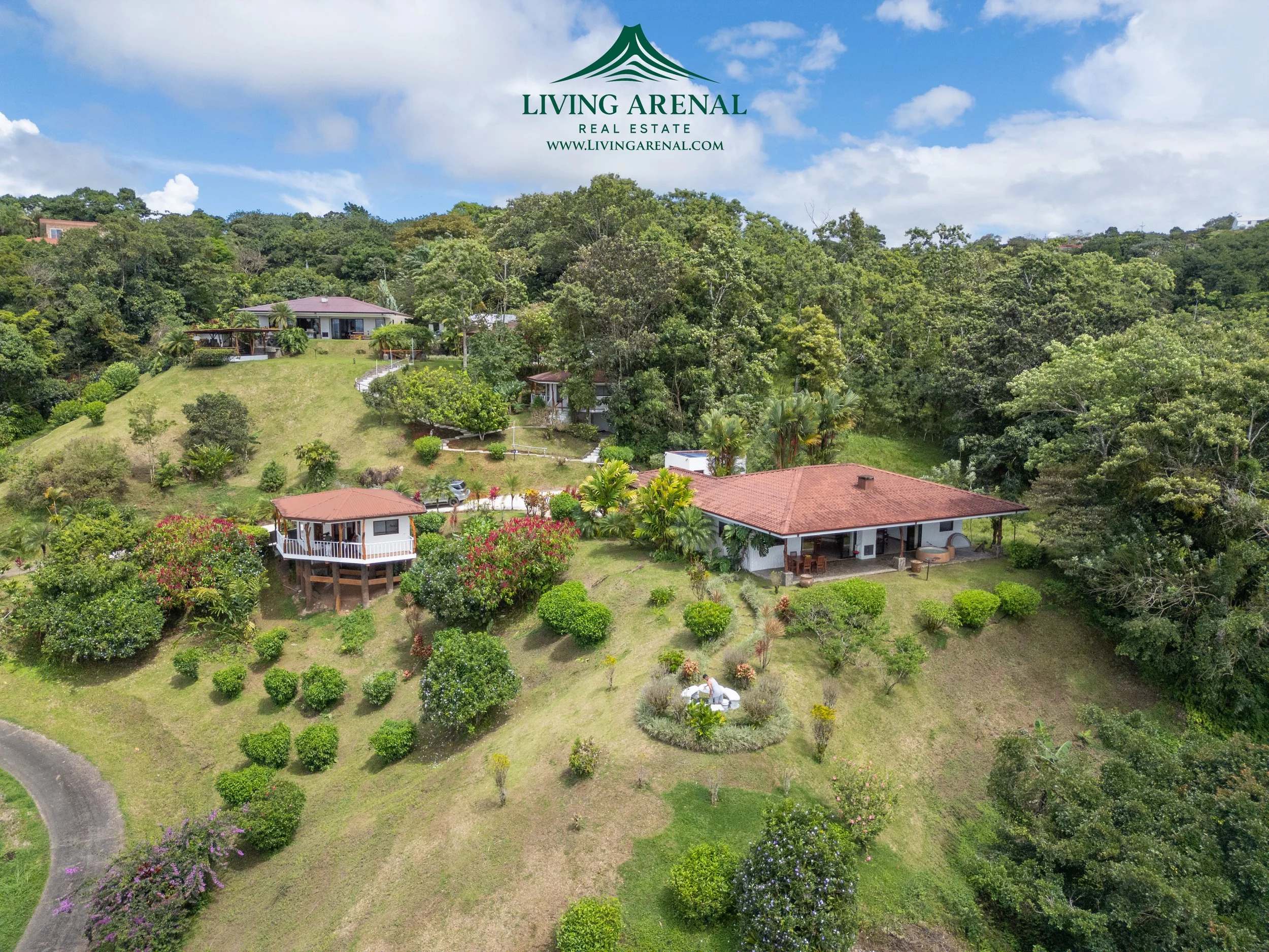 Aerial view of a residential area with multiple houses on a lush, green hillside, surrounded by trees and bushes, with a partly cloudy sky overhead.
