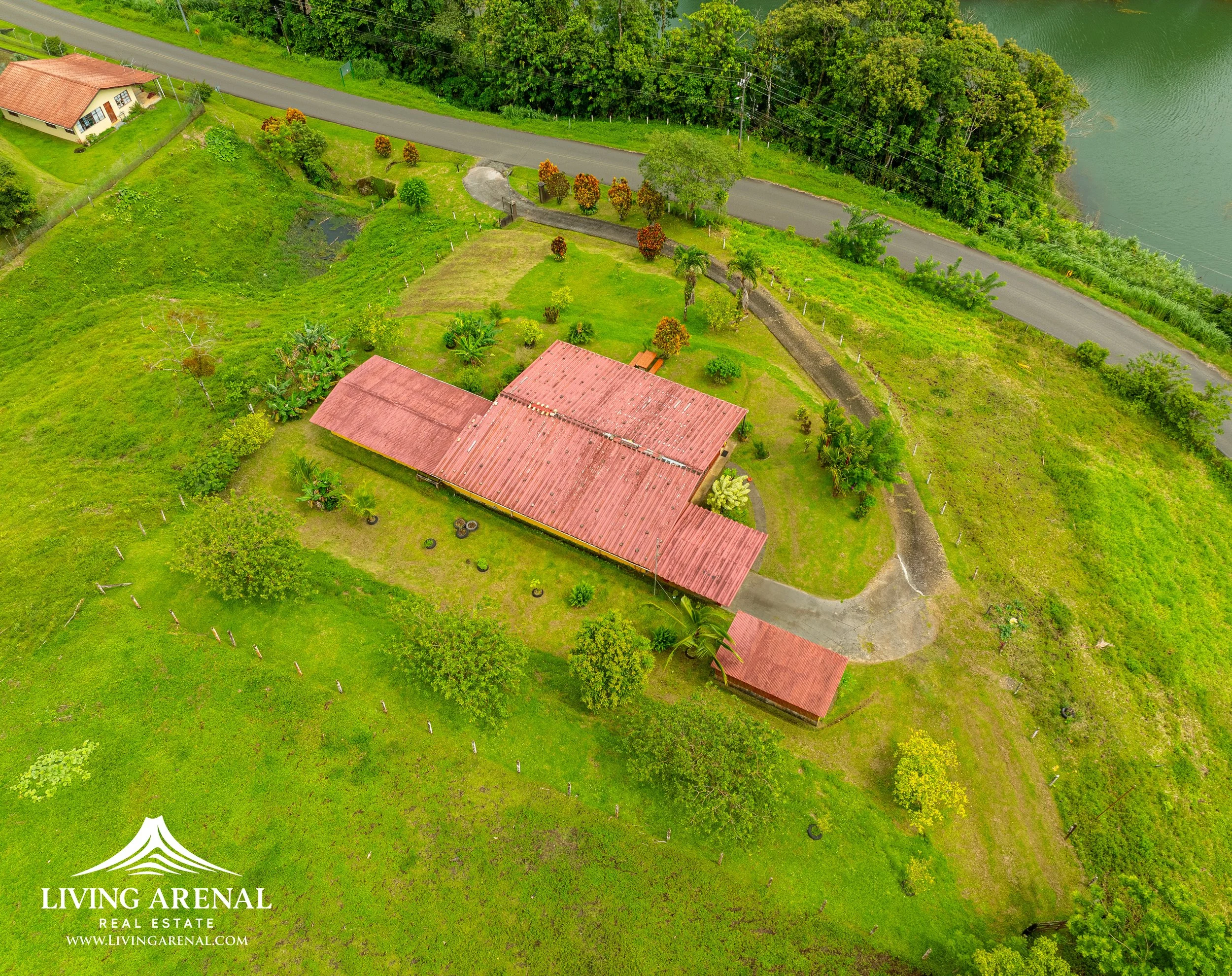Aerial view of a red-roofed house surrounded by green grass, trees, and a pond, with a nearby road and dense forest in the background.