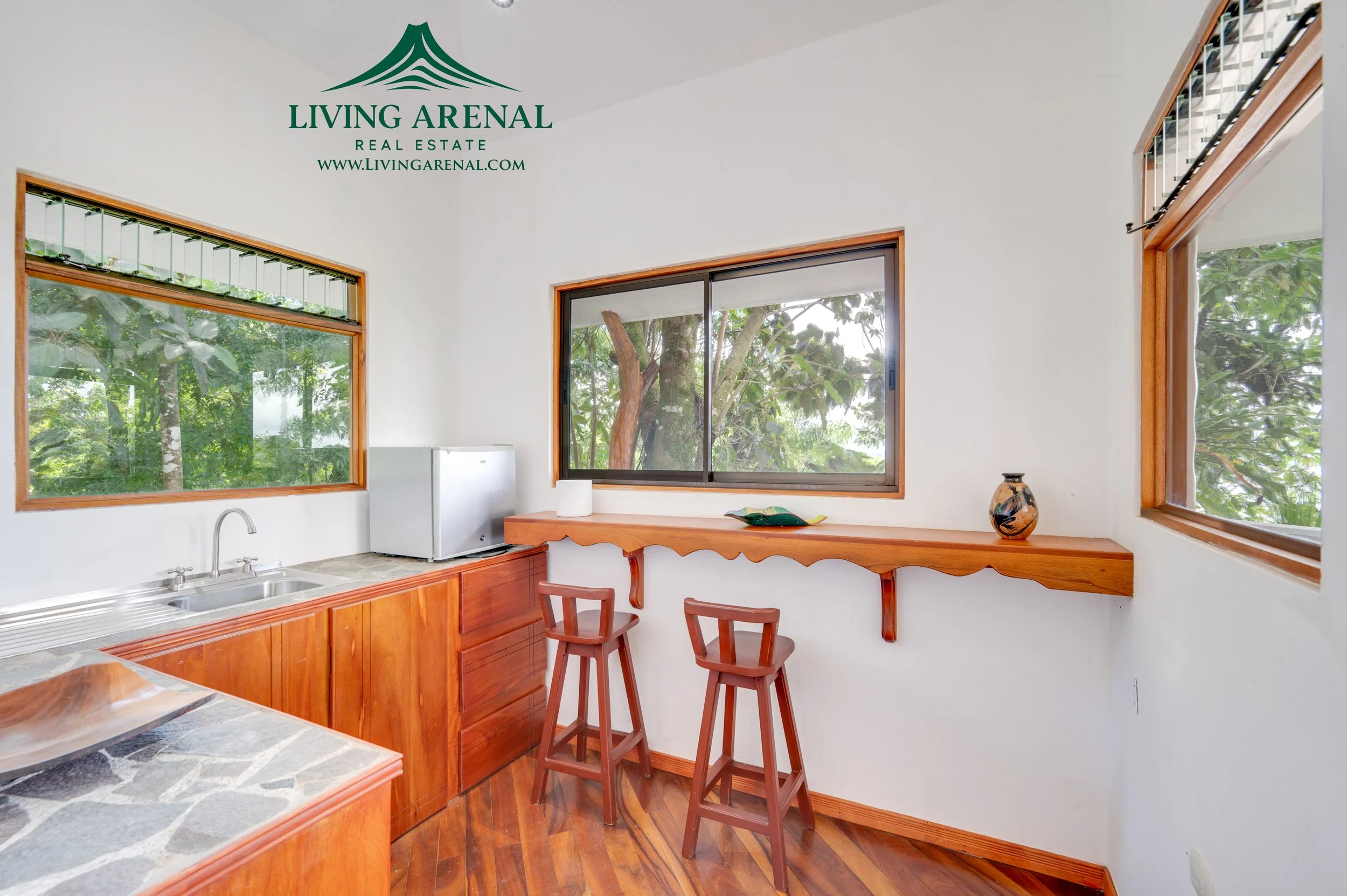 Kitchen with wooden cabinets, a window with a view of trees, a small refrigerator, a stone countertop, and a breakfast bar with two wooden stools.