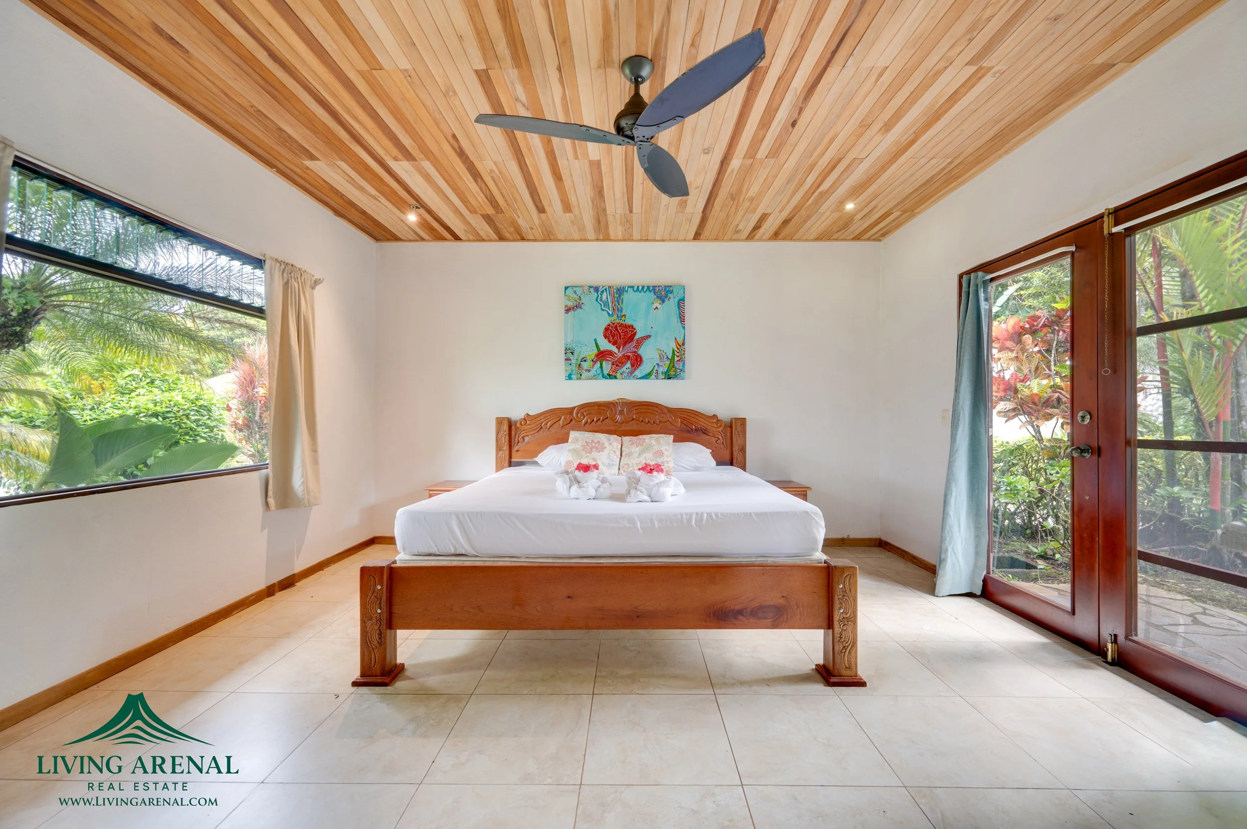 A bedroom with a wooden bed, white bedding, two folded towel swans, a colorful painting above the headboard, large windows with greenery outside, and a sliding glass door to the right with a view of plants. Ceiling fans and wooden ceiling panels are