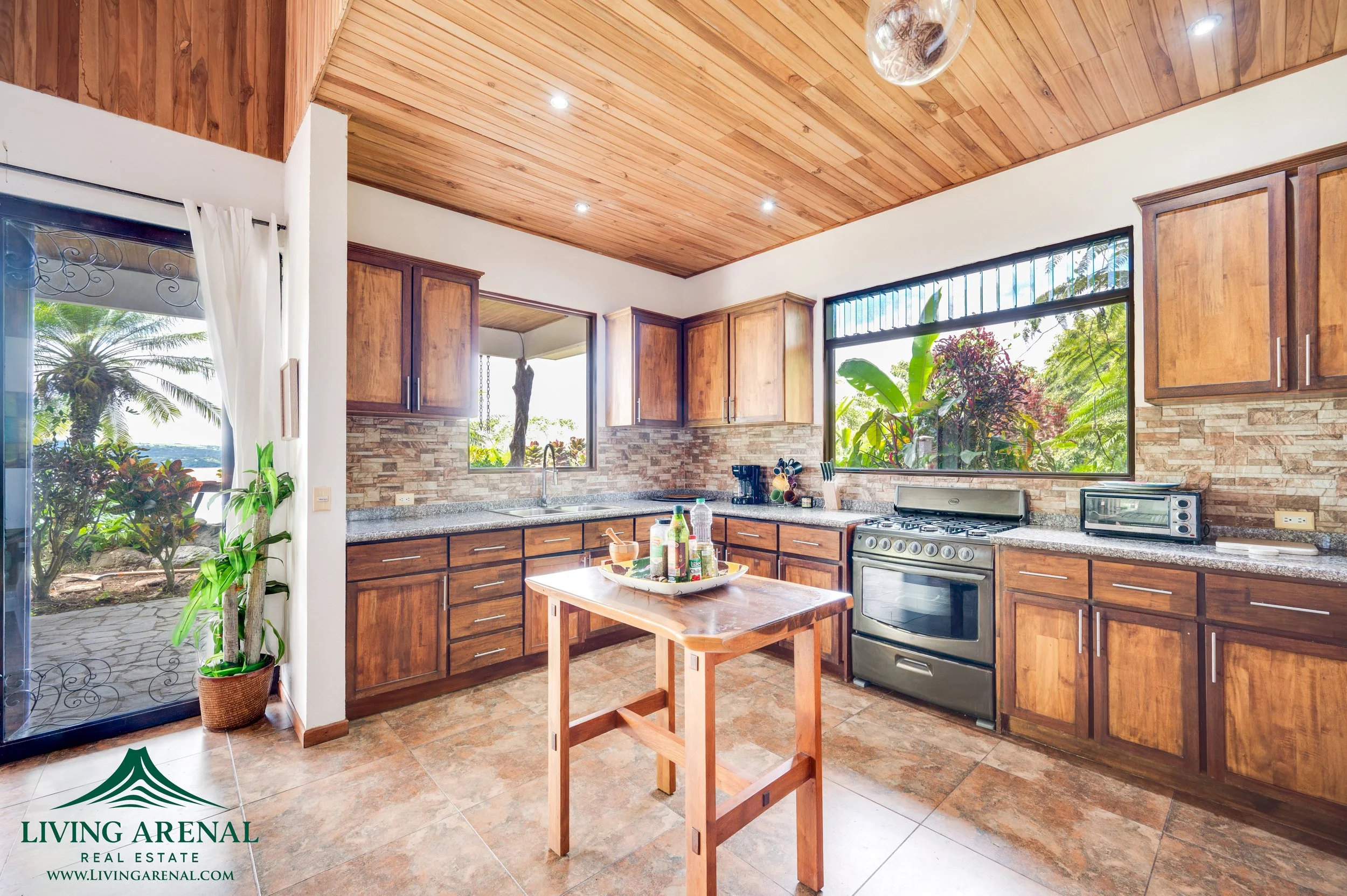 Spacious kitchen with wooden cabinets, granite countertops, a central wooden island, large windows showing greenery outside, stainless steel appliances, a potted plant near sliding door, and a tiled floor.