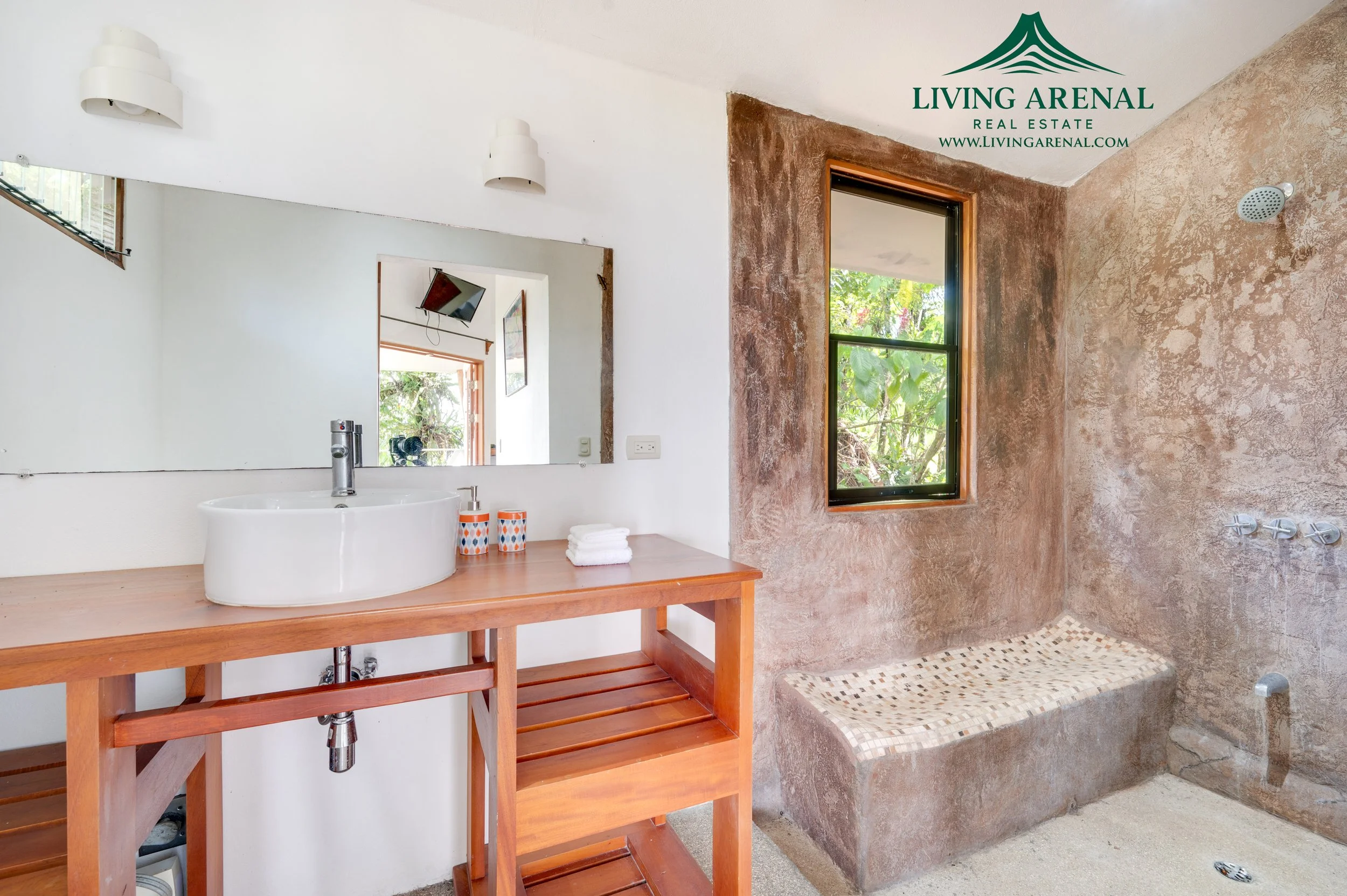 Bathroom with a wooden vanity, white vessel sink, large mirror, window with greenery, and shower area with built-in bench and stone walls.
