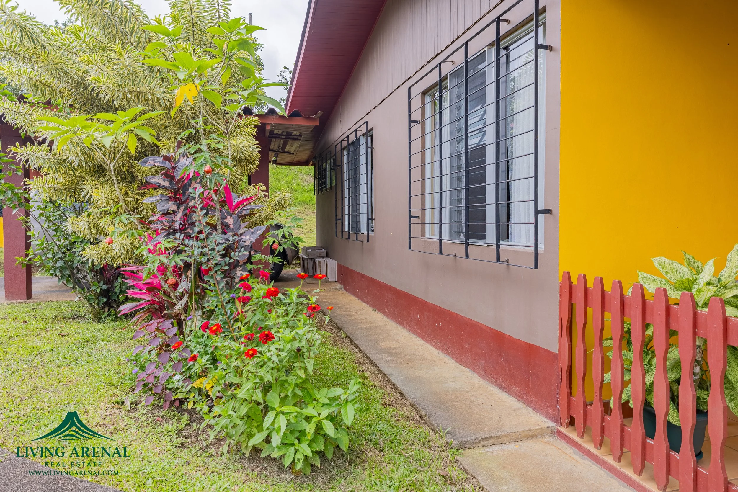 Side view of a house with three windows protected by black metal bars, a yellow and beige painted wall, a concrete pathway, a small red wooden fence, green grass, and assorted plants and shrubs.