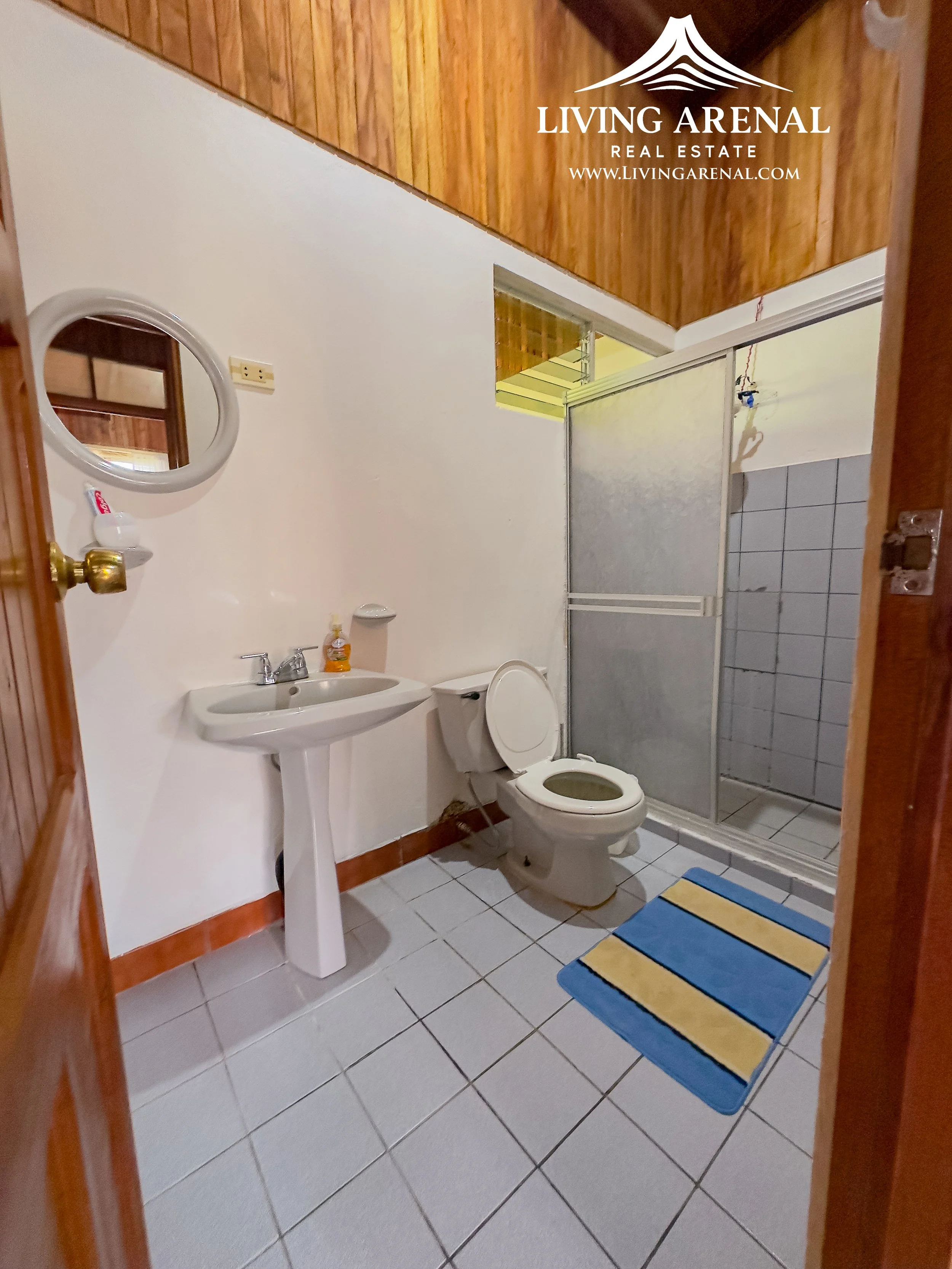 Bathroom with white tiled floor, white pedastal sink, toilet, small mirror, shower with sliding doors, and a blue and yellow striped rug.