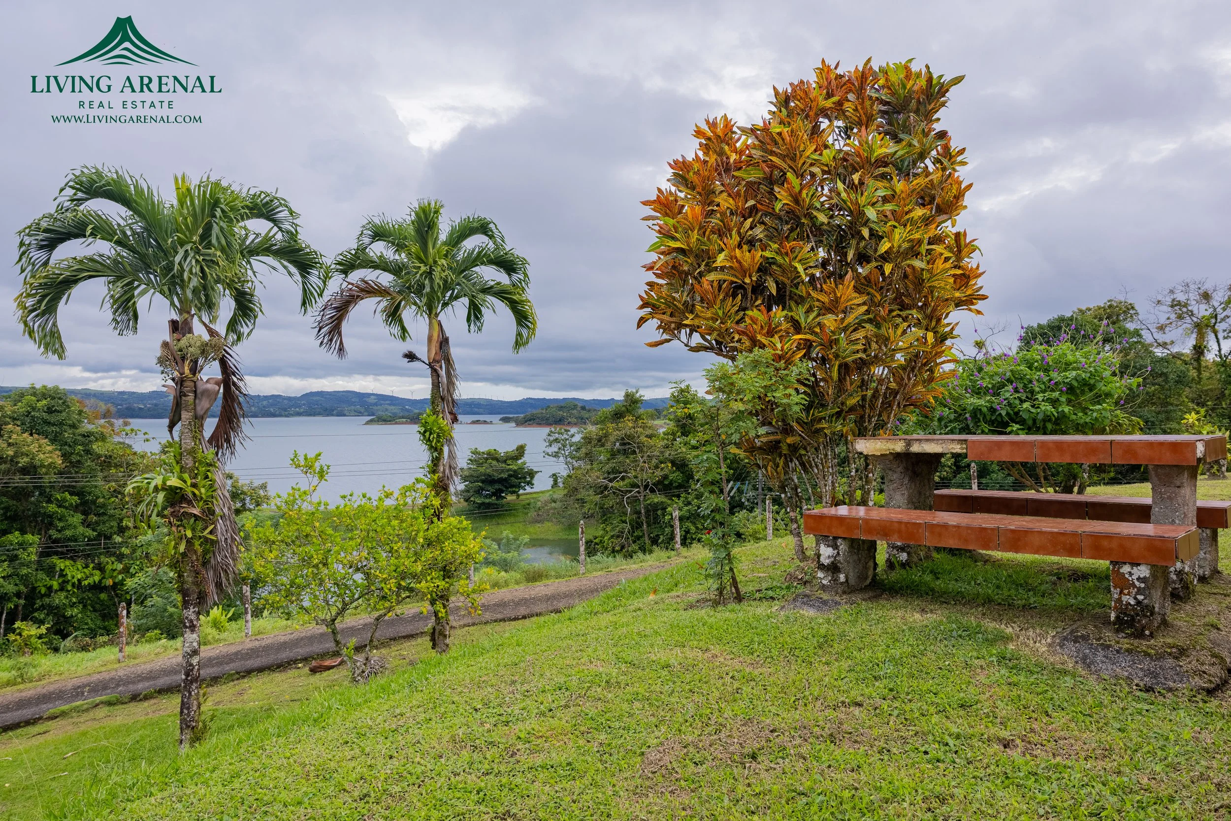 A scenic viewpoint with a wooden bench on stone supports, lush green grass, colorful tropical trees, and a large body of water with hills in the background. The sky is cloudy, and a logo for Living Arenal Real Estate is in the top left corner.