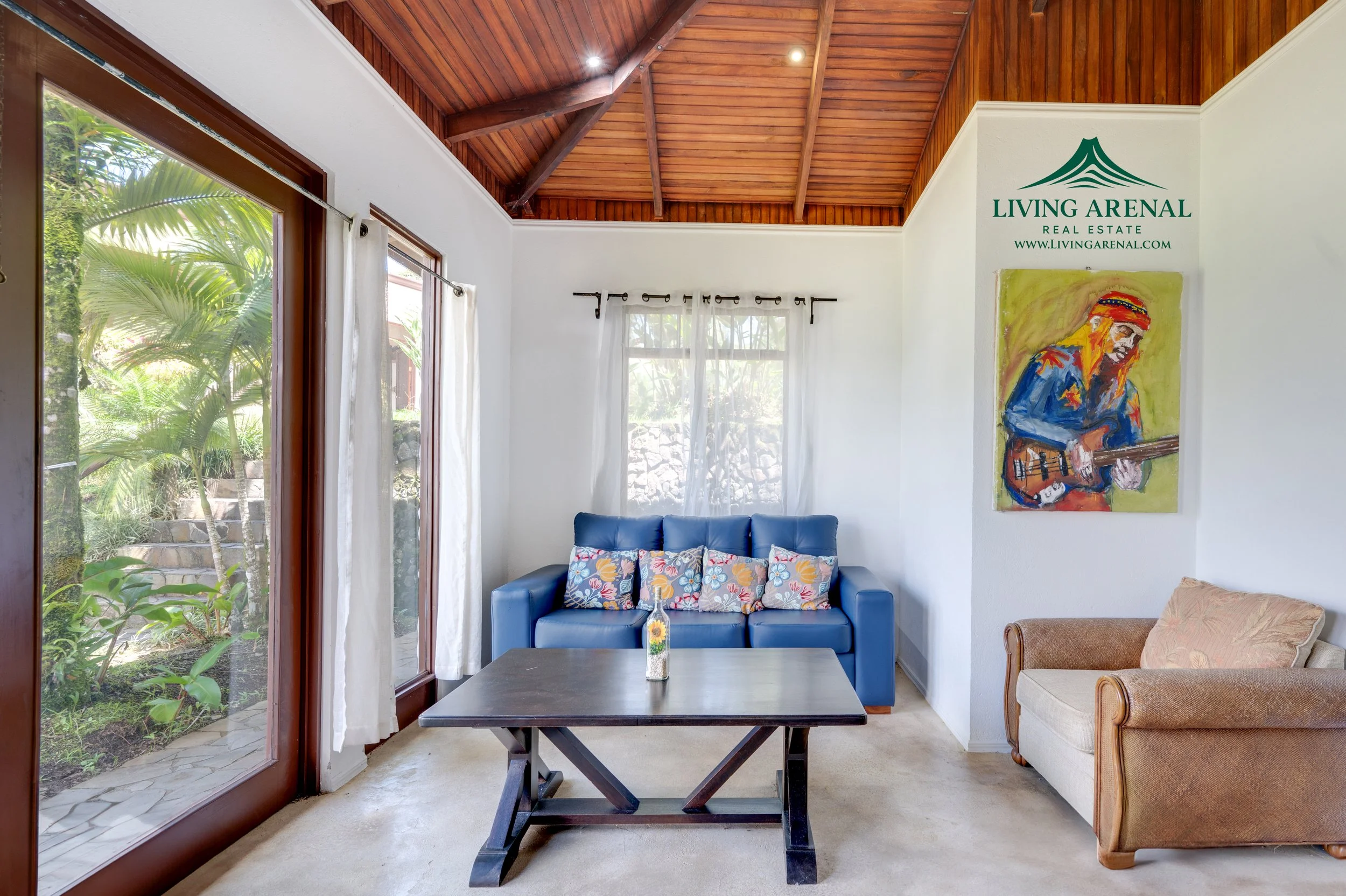 Bright living room with a wooden vaulted ceiling, glass sliding door to outside greenery, white walls, a blue sofa with colorful patterned pillows, an orange armchair, a dark wood coffee table, a vase with flowers, and artwork on the wall including a