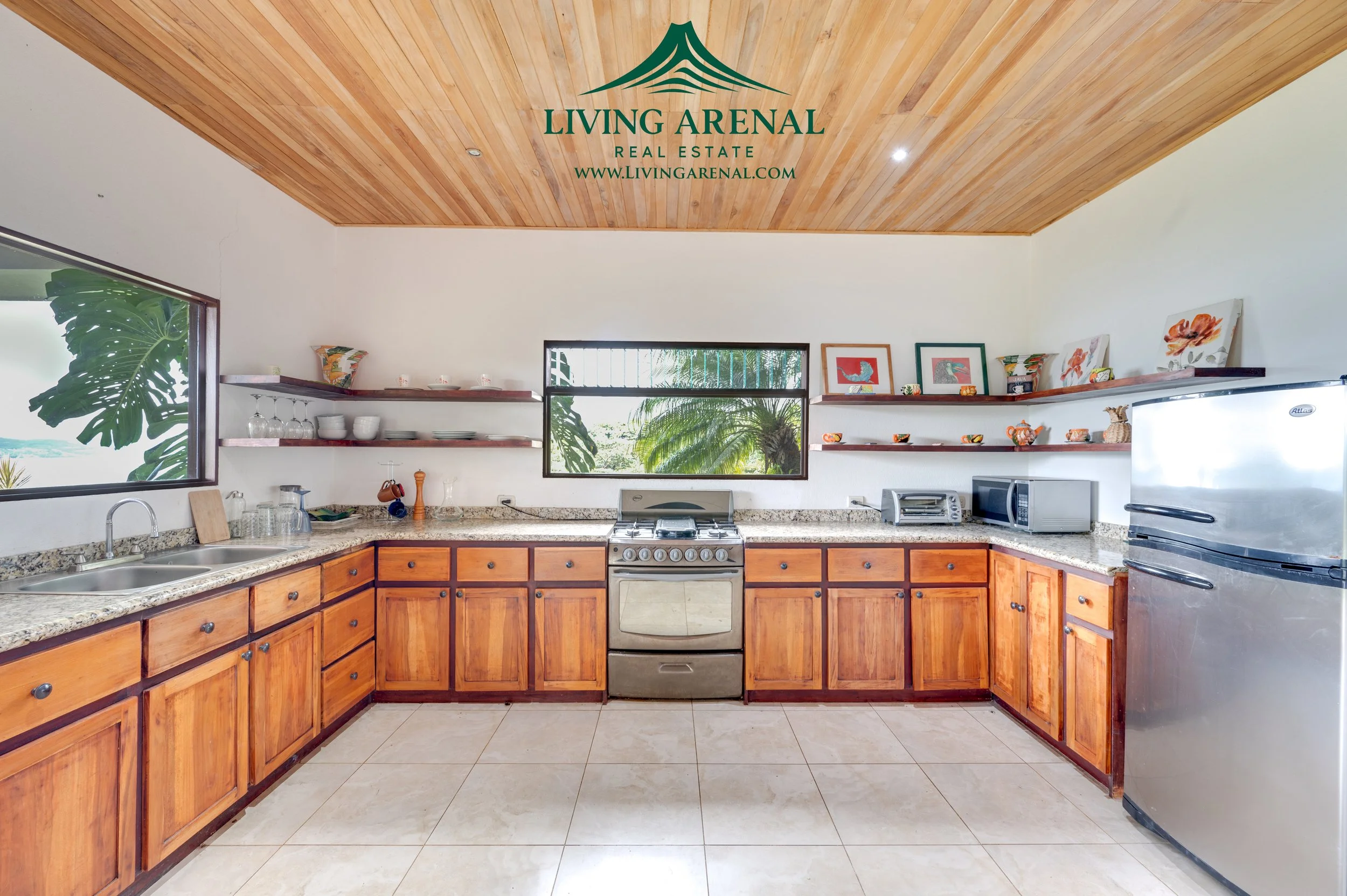 Kitchen with wooden cabinets, granite countertops, stainless steel stove, microwave, toaster, and fridge, with open shelves displaying dishes and decorative items, window showing leafy tropical greenery.