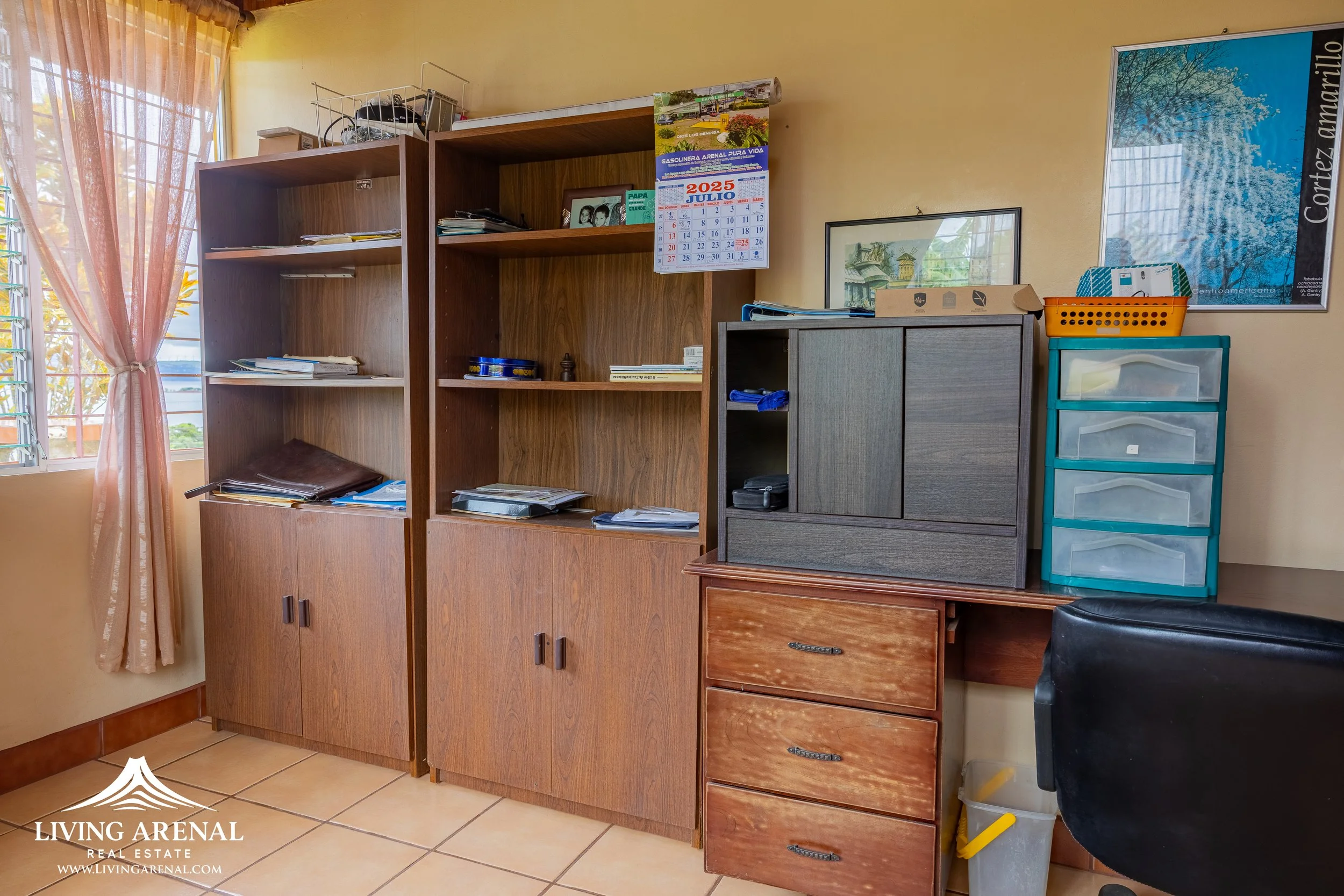Office desk with storage shelves and drawers, calendar on wall, and a large window with pink curtains.