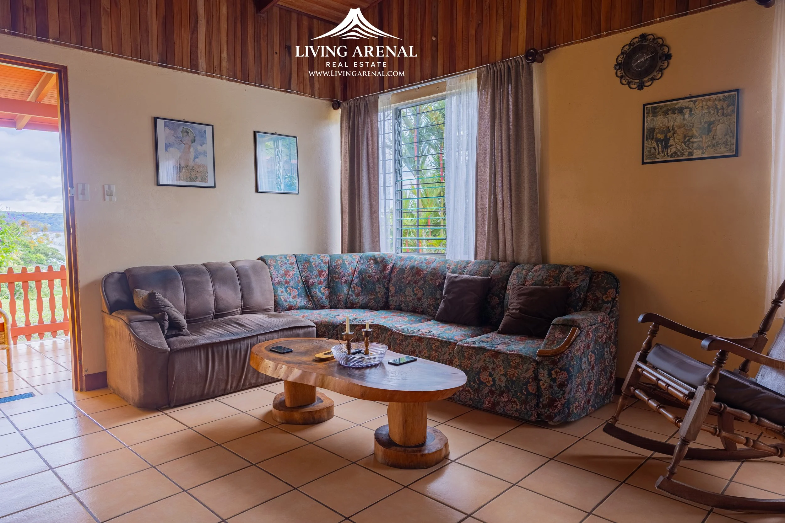 Living room with a tiled floor, two couches, and a wooden coffee table. One couch is leather, and the other has a floral pattern. There are four pillows on the couches. A rocking chair is next to the floral couch. A window with curtains lets in natur