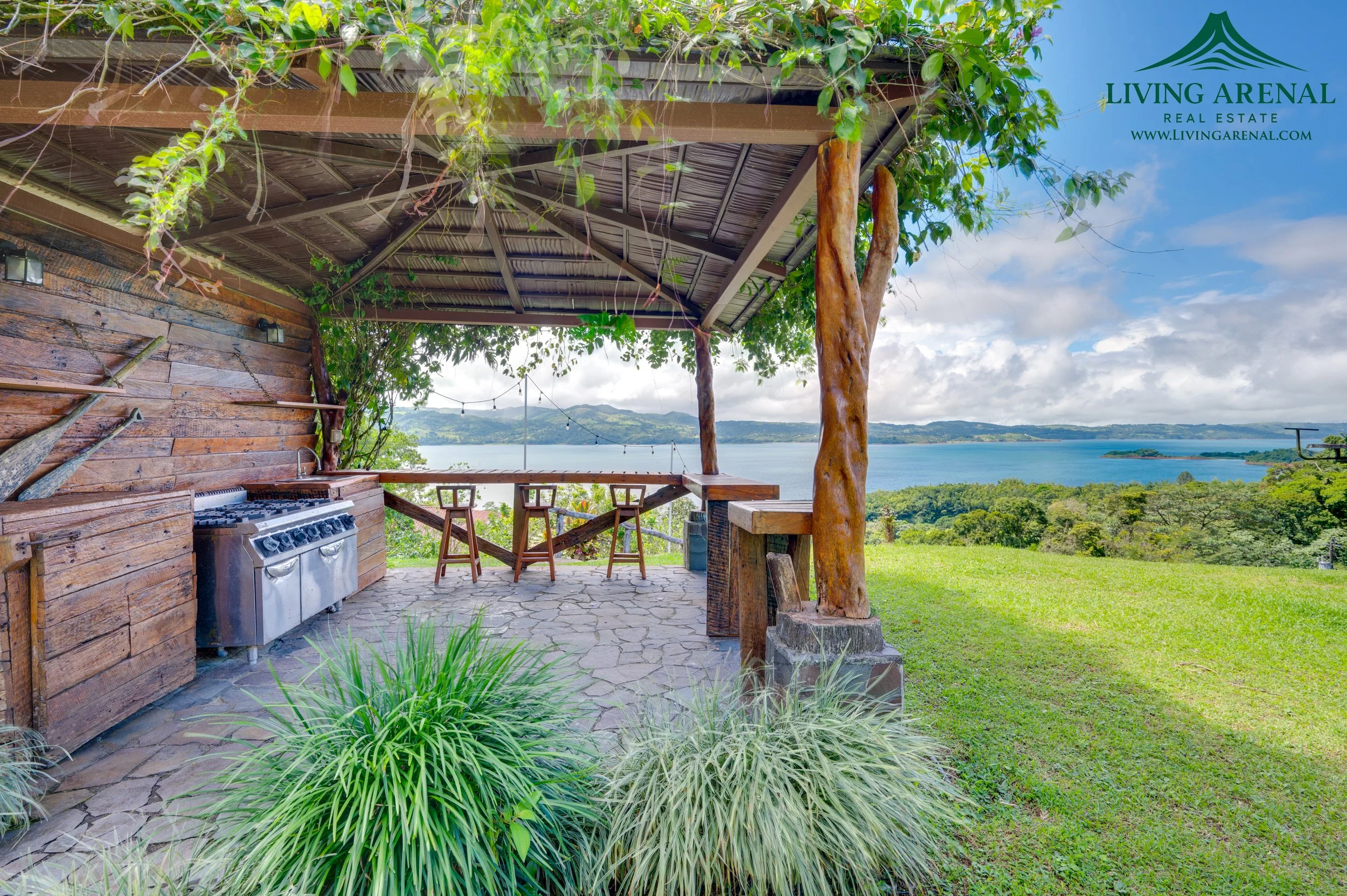 Outdoor patio with wooden bar, chairs, and a grill, overlooking a scenic lake and hills under a cloudy sky, surrounded by trees and greenery.