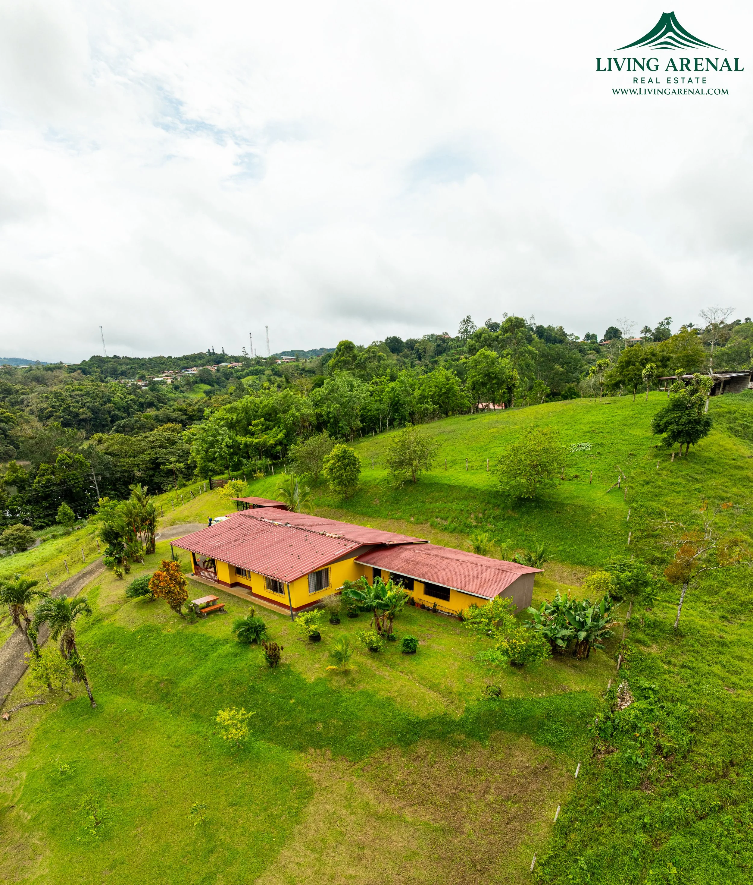 A yellow house with a red roof is situated on a lush green hillside, with trees and plants surrounding it under a cloudy sky.