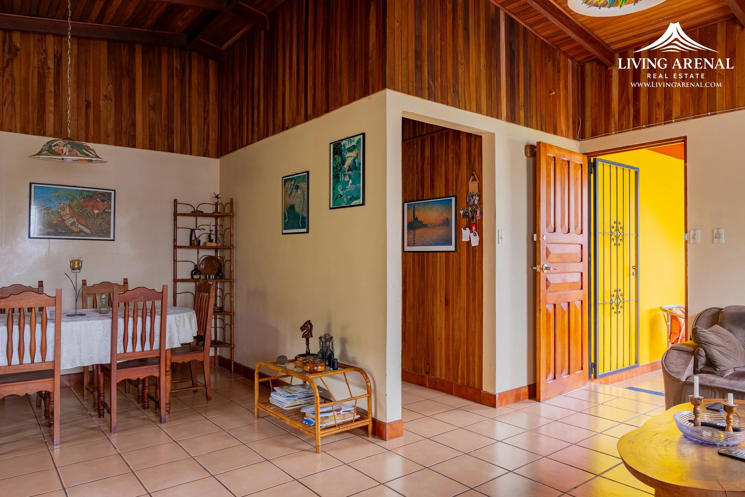 Living room with a wooden ceiling and tile floor, showing a dining area with a white tablecloth, wooden chairs, a bookshelf, framed artwork on the walls, a wooden door, and an open yellow door leading outside.