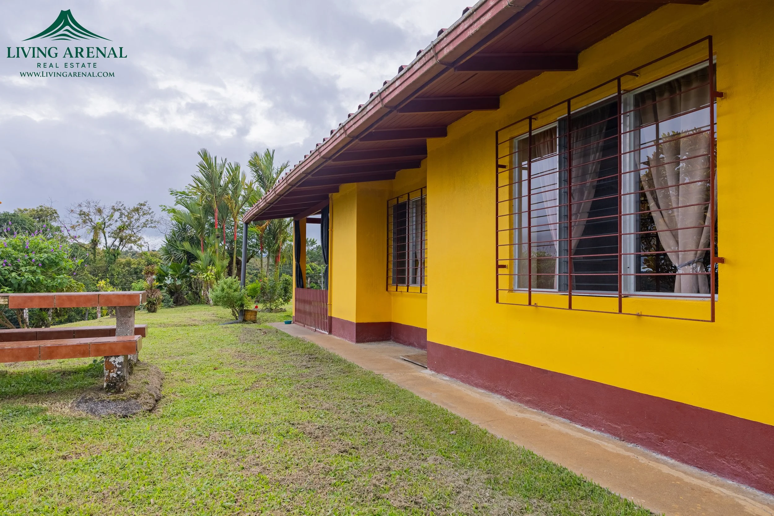 Yellow house with red window bars and a brown roof, surrounded by green grass, plants, and trees, under a cloudy sky.