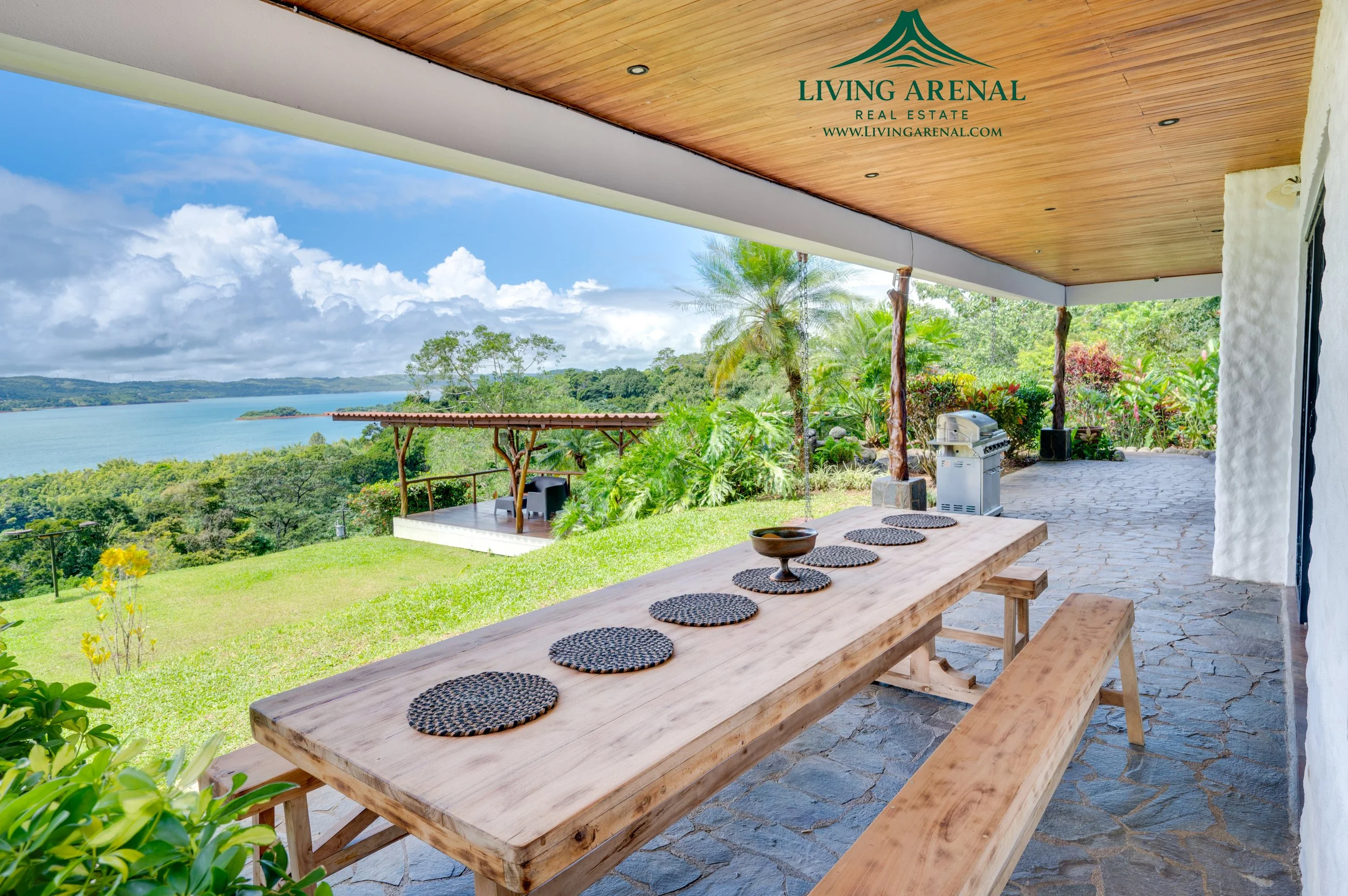 Covered outdoor patio with a wooden dining table and benches, surrounded by lush greenery and overlooking a large lake or river with islands, under a partly cloudy sky.
