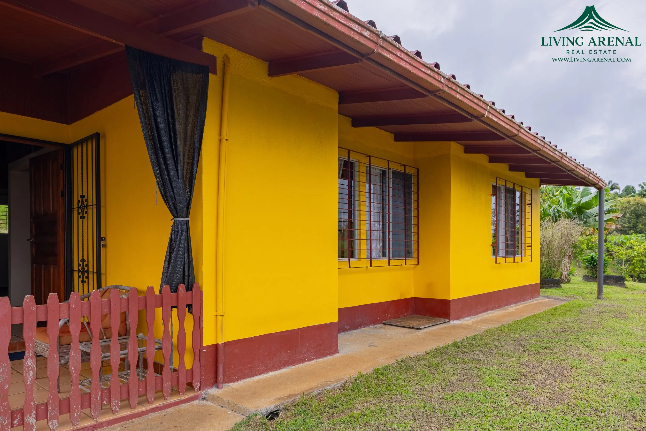 A yellow residential house with red trim and a small front porch with a red picket fence. The house has barred windows, a wooden door, and a brown tiled roof. There is a garden with green grass and plants on the right side.