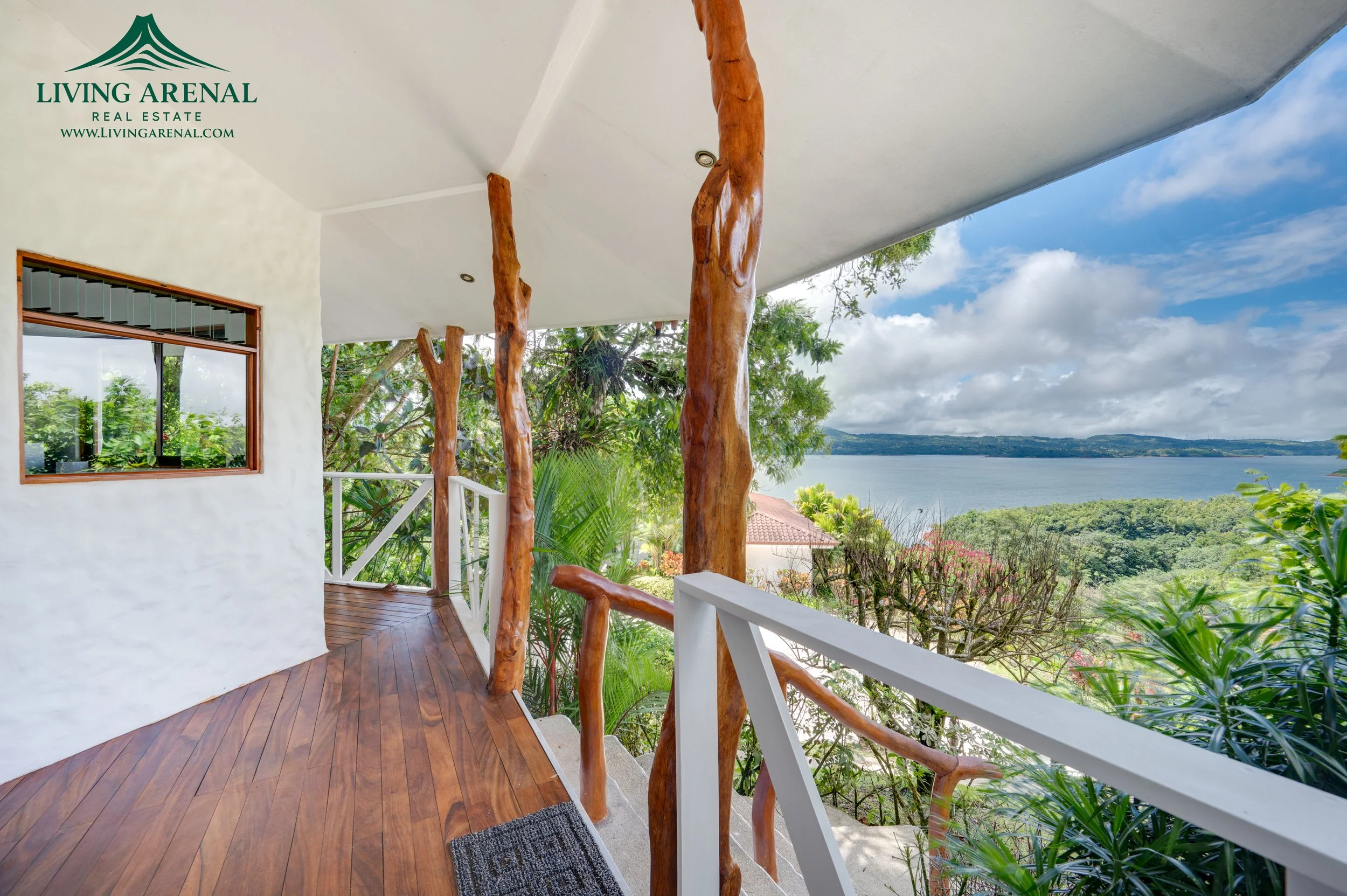 View from a balcony showing wooden posts, railing, lush greenery, and a lake with cloudy sky in the background.