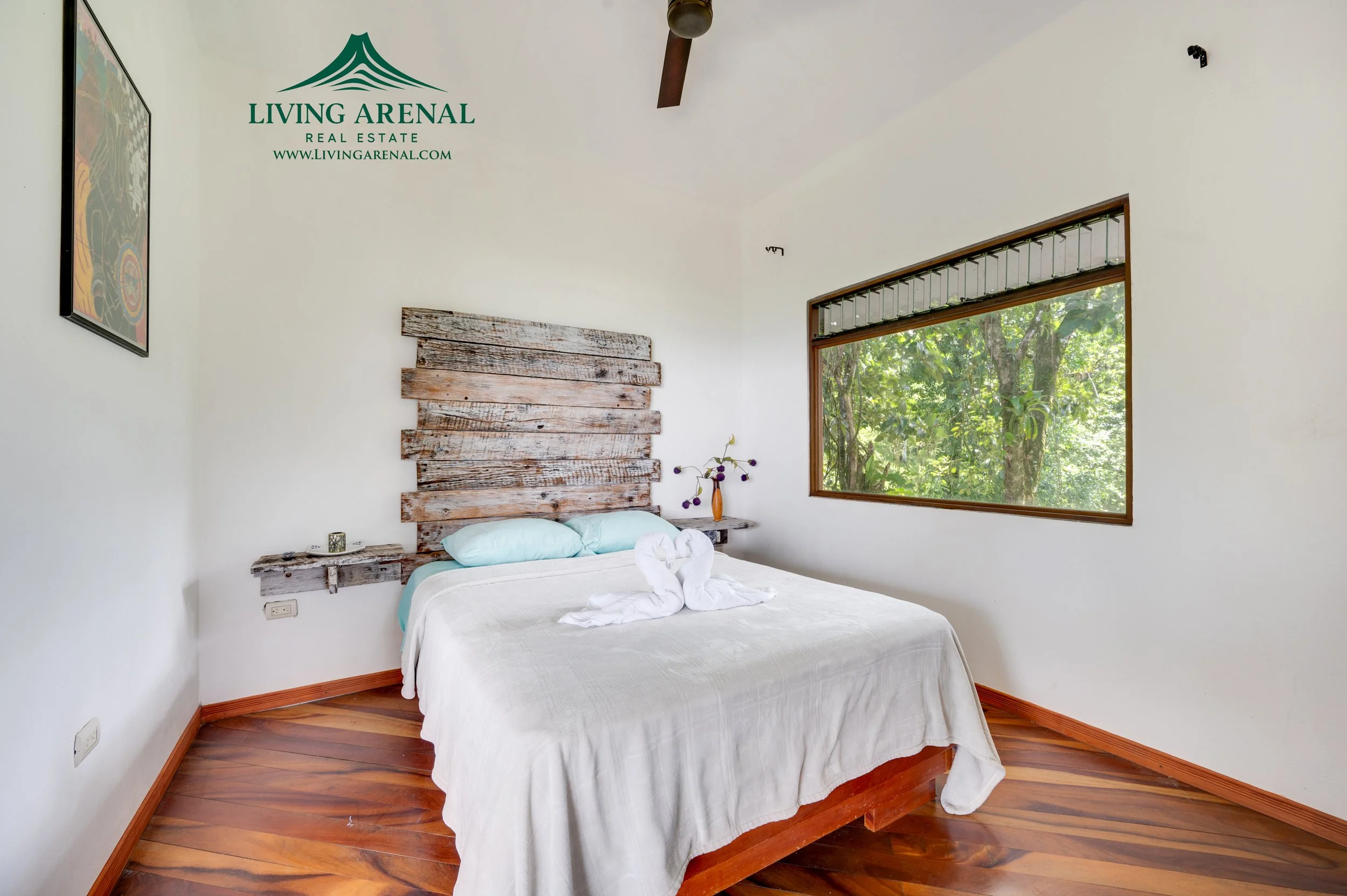 A minimalist bedroom with a white bed with pillows, towel swans, a rustic wooden headboard, a small shelf with a vase of flowers, a large window showcasing green trees outside, and wooden flooring. The room features a ceiling fan and wall decor that