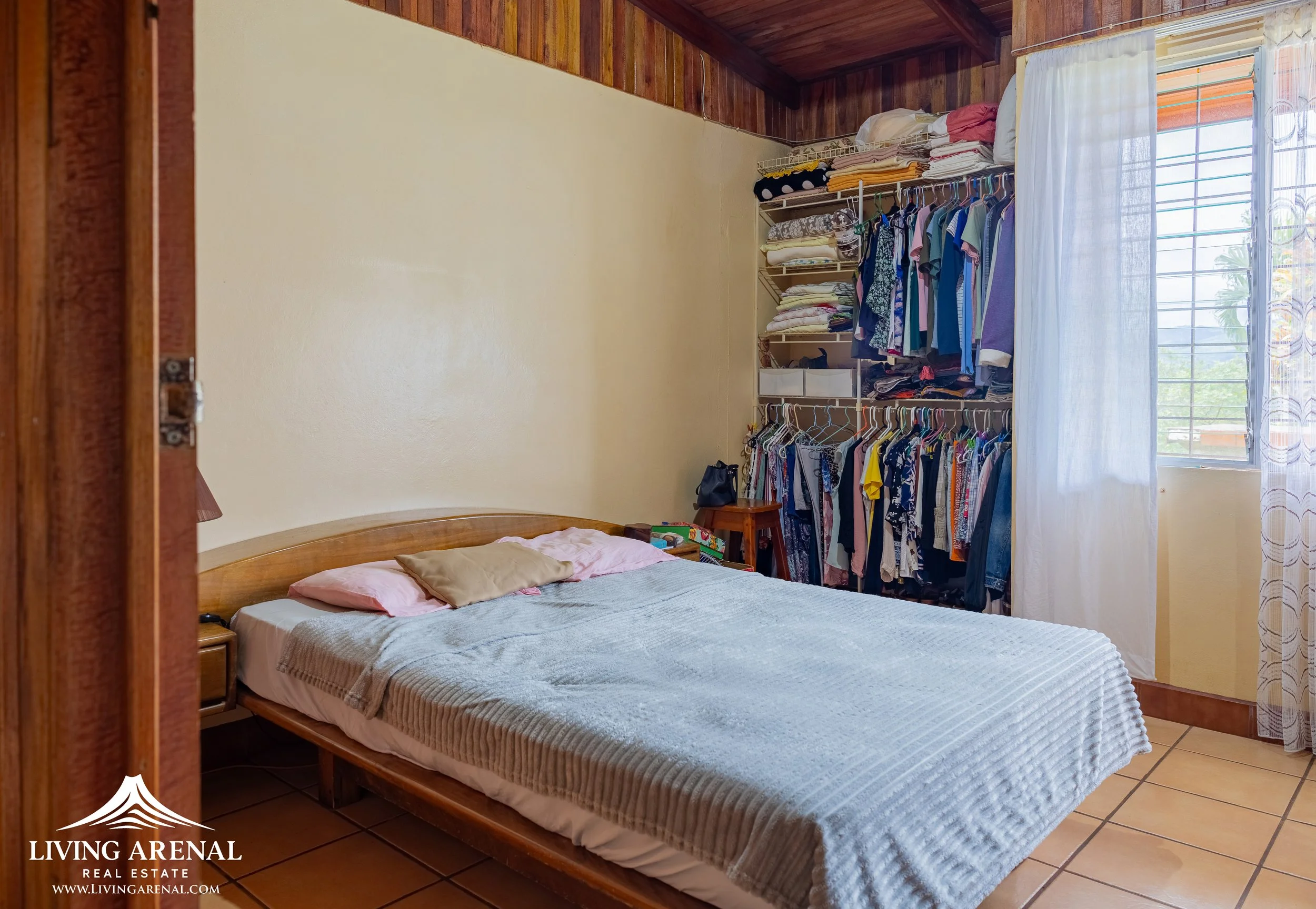 Bedroom with a wooden bed, multiple pillows, and a clothing rack filled with clothes near a window with white curtains.