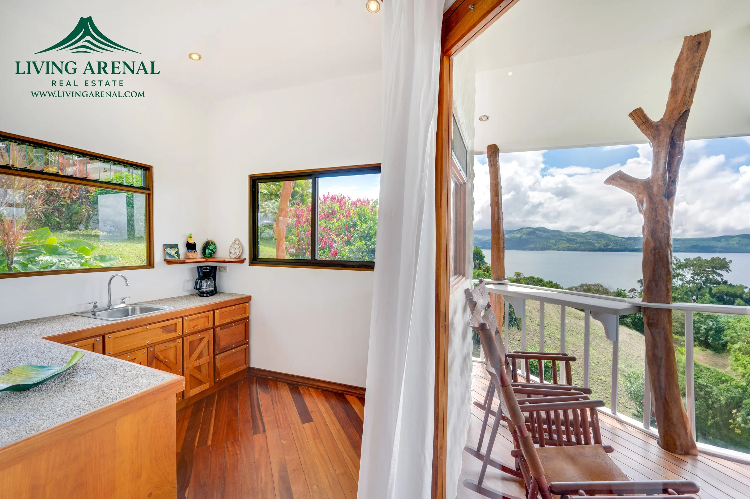 Interior view of a kitchen with wooden cabinets, a countertop with a sink, and two windows showing greenery outside. An open door leads to a balcony with wooden chairs, overlooking a lake and hills under a cloudy sky.