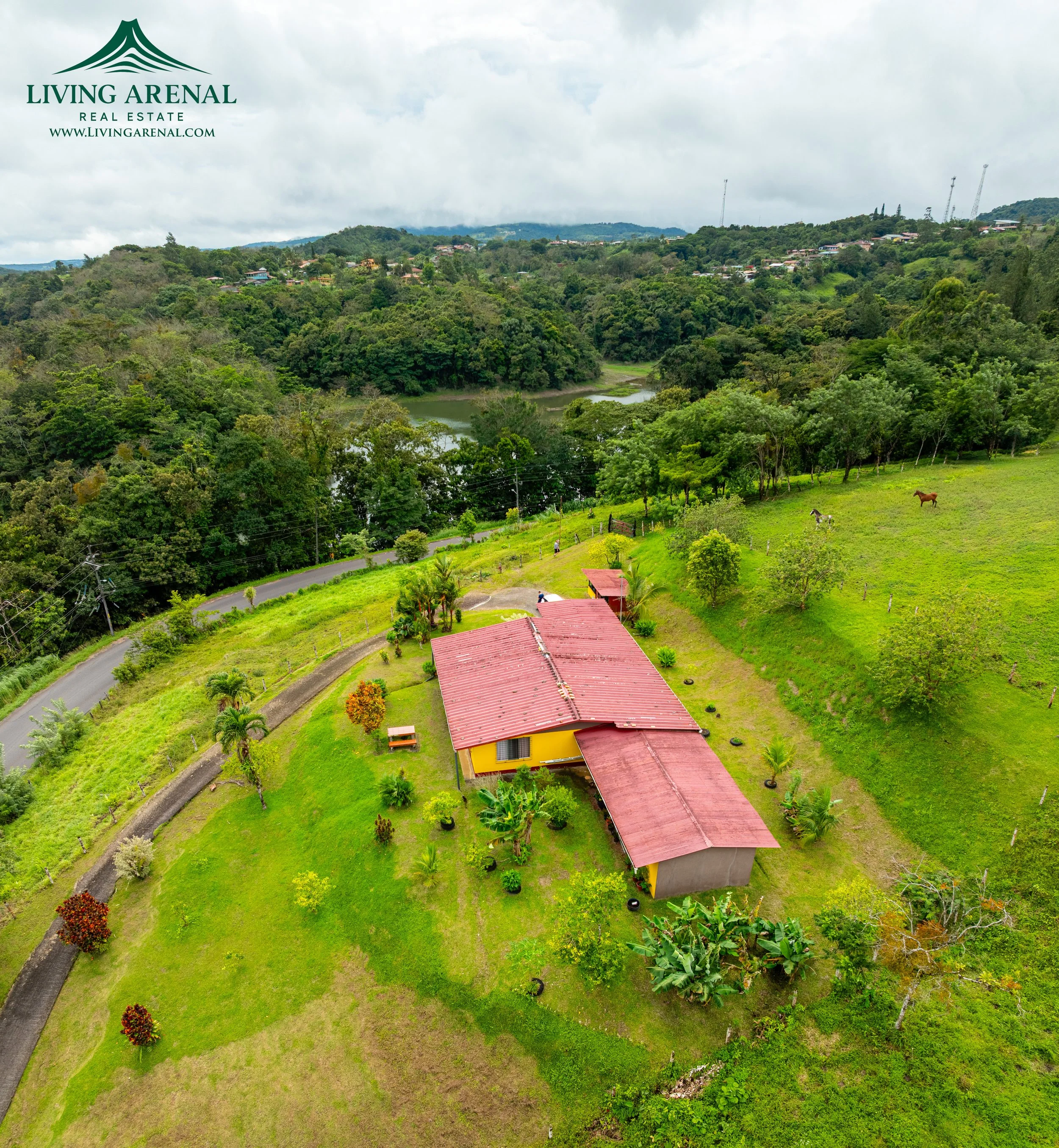 Aerial view of a countryside property with a yellow house and red roof, surrounded by lush greenery, a winding road, and horses grazing on a hill.