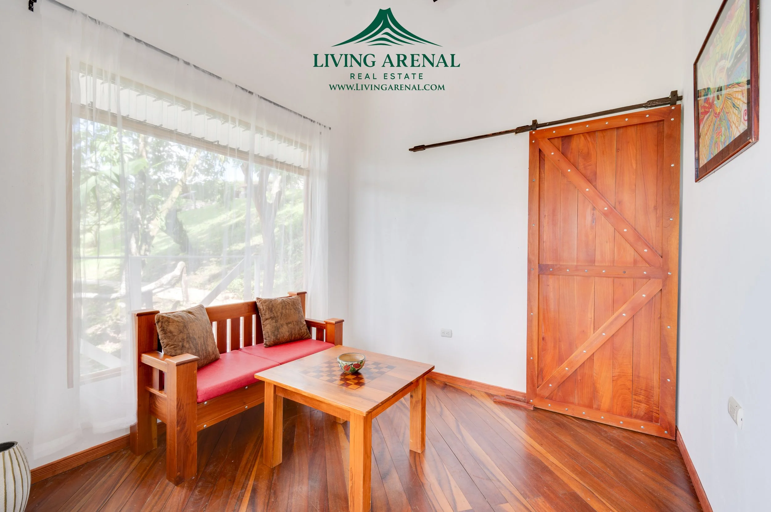 A cozy room featuring wooden furniture including a bench with cushions, a small coffee table with a decorative bowl, a large window with sheer curtains revealing greenery outside, and a sliding wooden barn door. The floor and furniture are made of wa