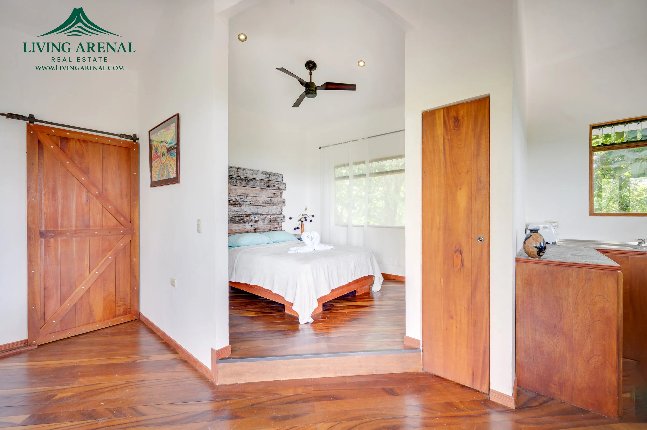 Interior of a bedroom with wooden floors, a bed with white linens, a rustic wood headboard, a small kitchen area with a sink, a window with view of greenery, and wooden doors.