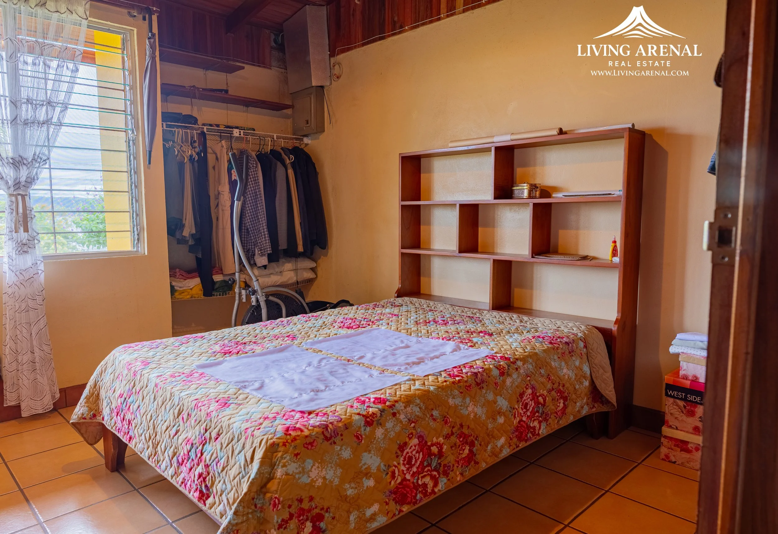 A small bedroom with a bed covered with a patterned quilt and two white towels on top. To the left, there's a window with curtains, and an open closet with hanging clothes, a shelf, and a vacuum cleaner. To the right, a wooden shelf is attached to th