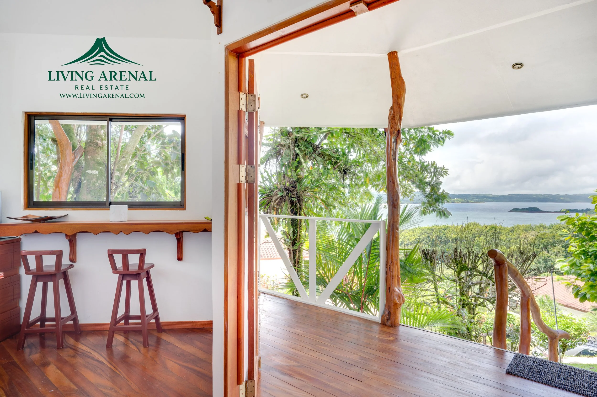 Balcony with wooden railing and flooring overlooking lush green trees, a body of water, and cloudy sky.
