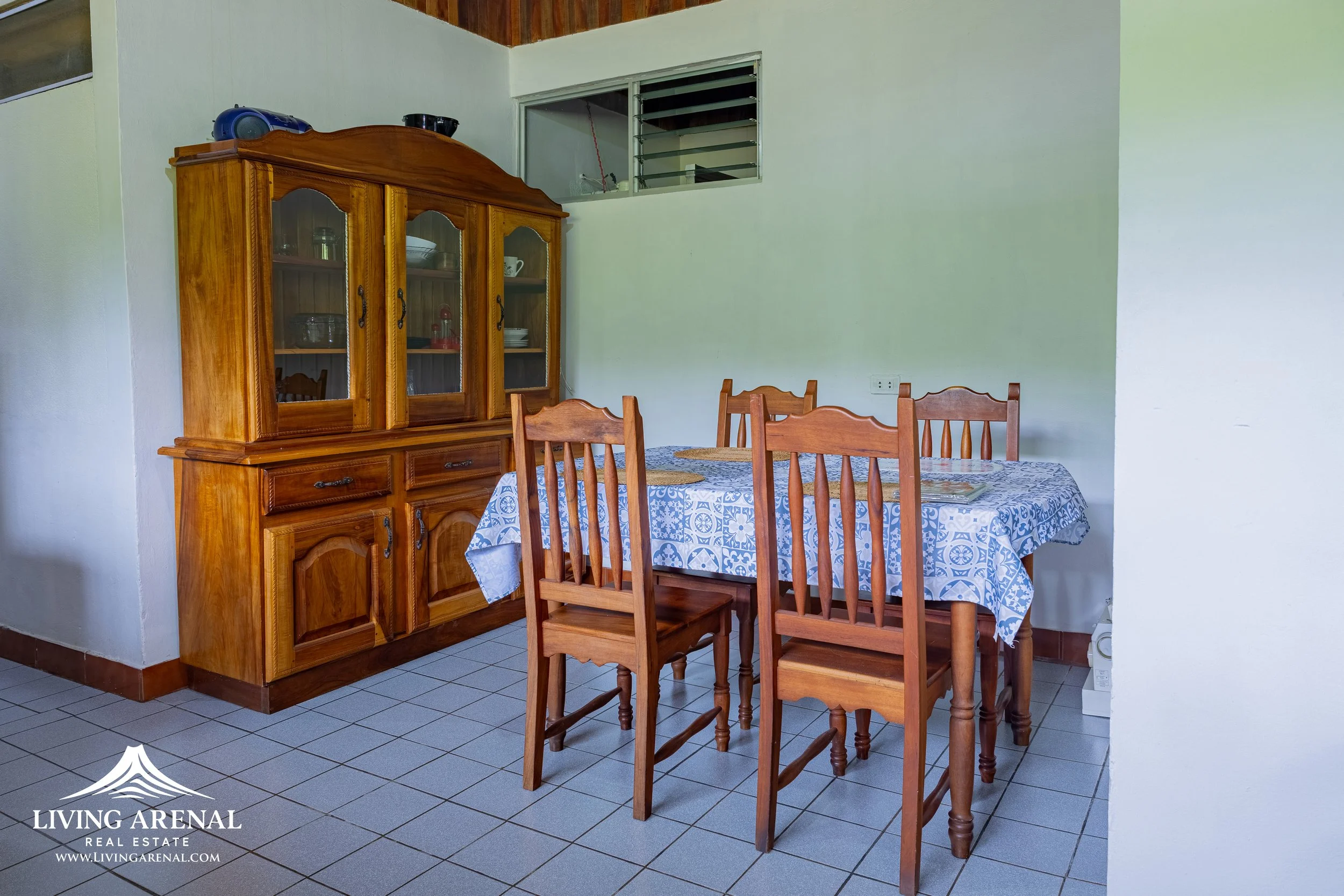 A dining room with a wooden dining table covered with a blue and white patterned tablecloth, surrounded by six matching wooden chairs. A wooden china cabinet with glass doors displays dishes and glassware, positioned against a pale green wall. There