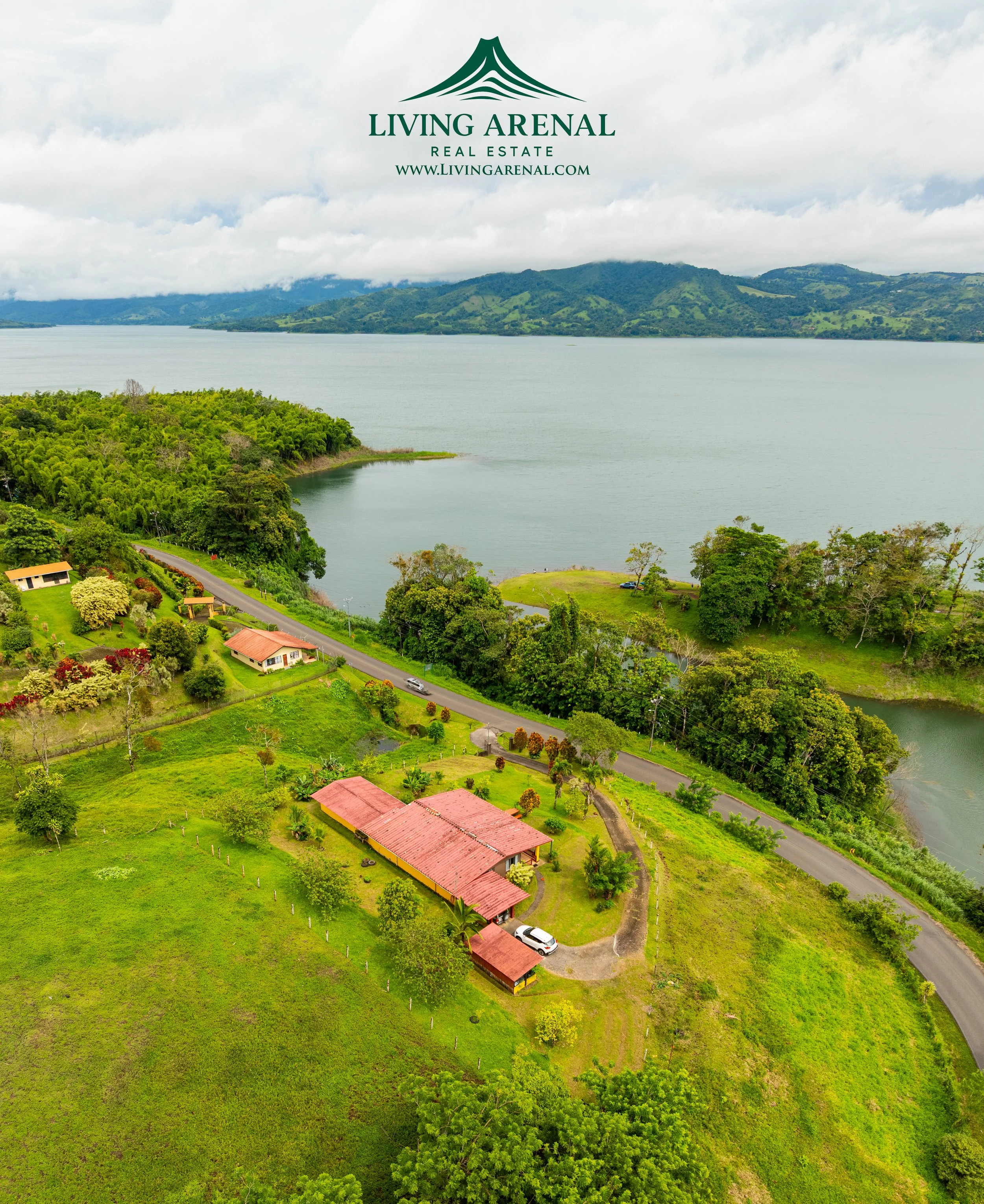 Aerial view of a lakeside property with green lawns, trees, and a red-roofed house next to a curved road, overlooking a large lake with hills in the background.