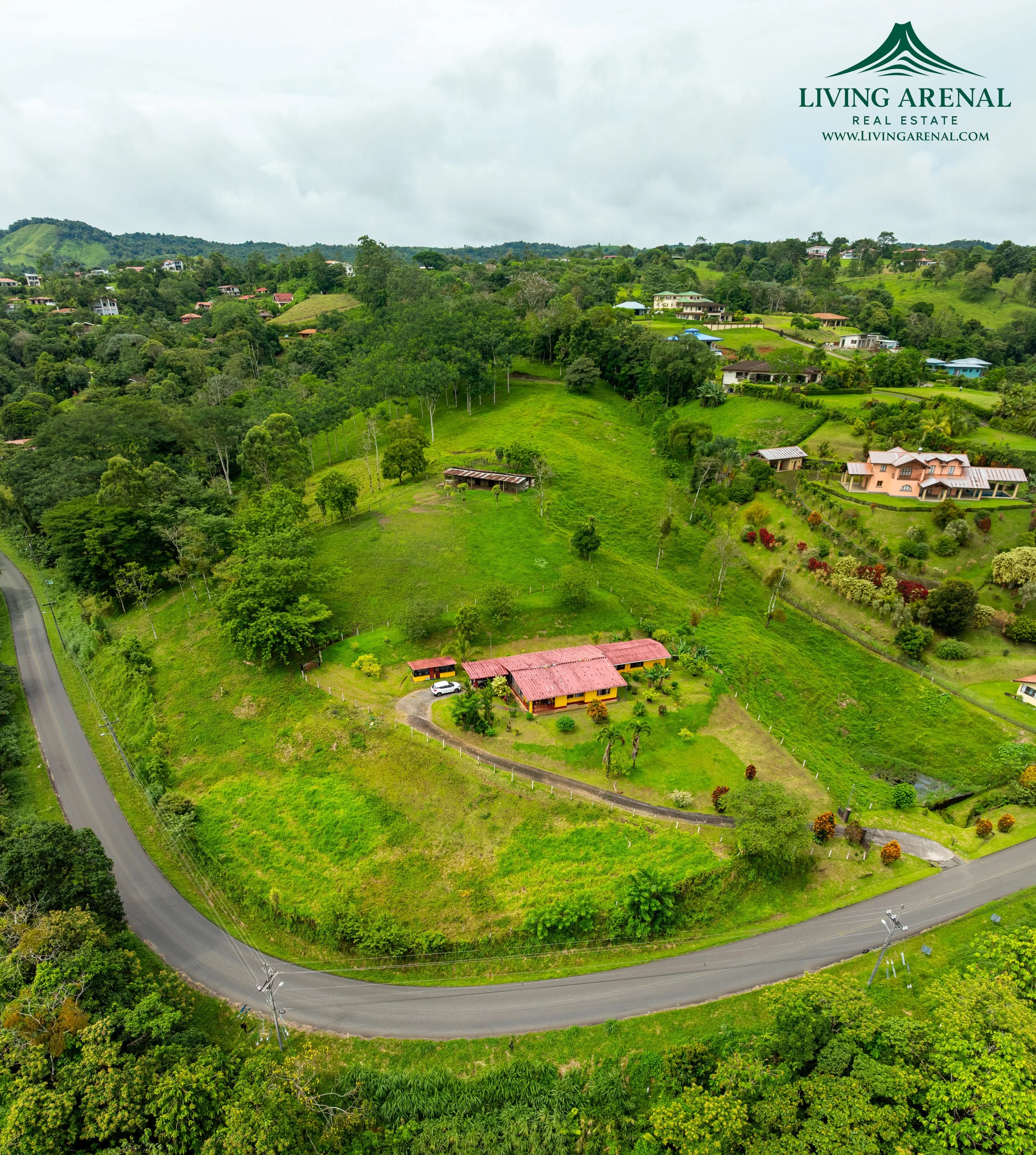 Aerial view of a green hilly area with several houses, roads, and trees, with the logo and website of Living Arenal Real Estate in the top right corner.