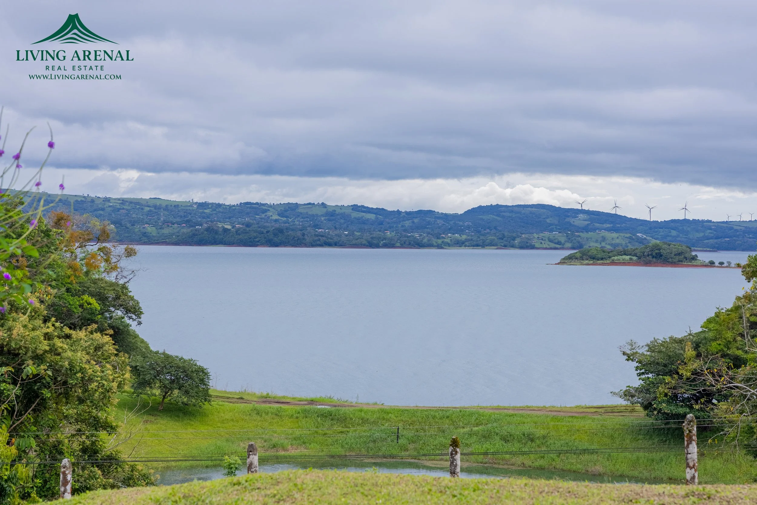 A scenic view of a large body of water with green hills and wind turbines in the background, cloudy sky, and trees in the foreground, with a "Living Arenal" real estate logo on the top left corner.