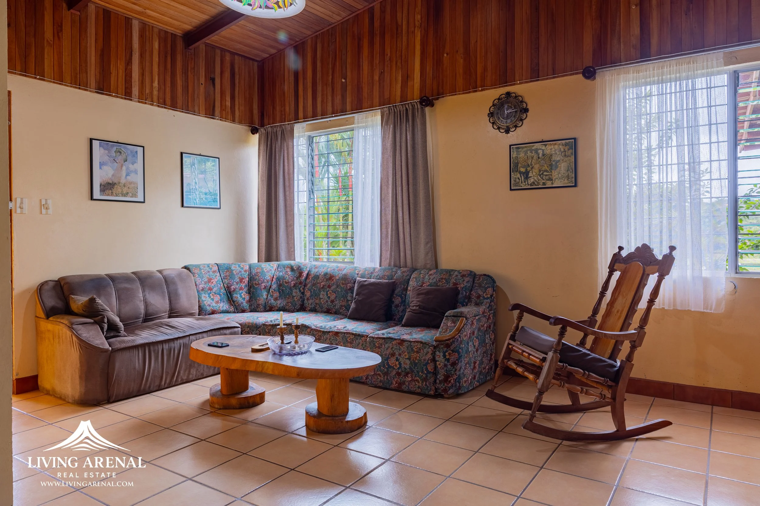 Living room with tiled floor, wooden ceiling, and yellow walls. Contains a sectional sofa with floral and brown upholstery, a wooden coffee table with candlesticks and remote controls, a wooden rocking chair, and framed pictures on the wall. Large wi