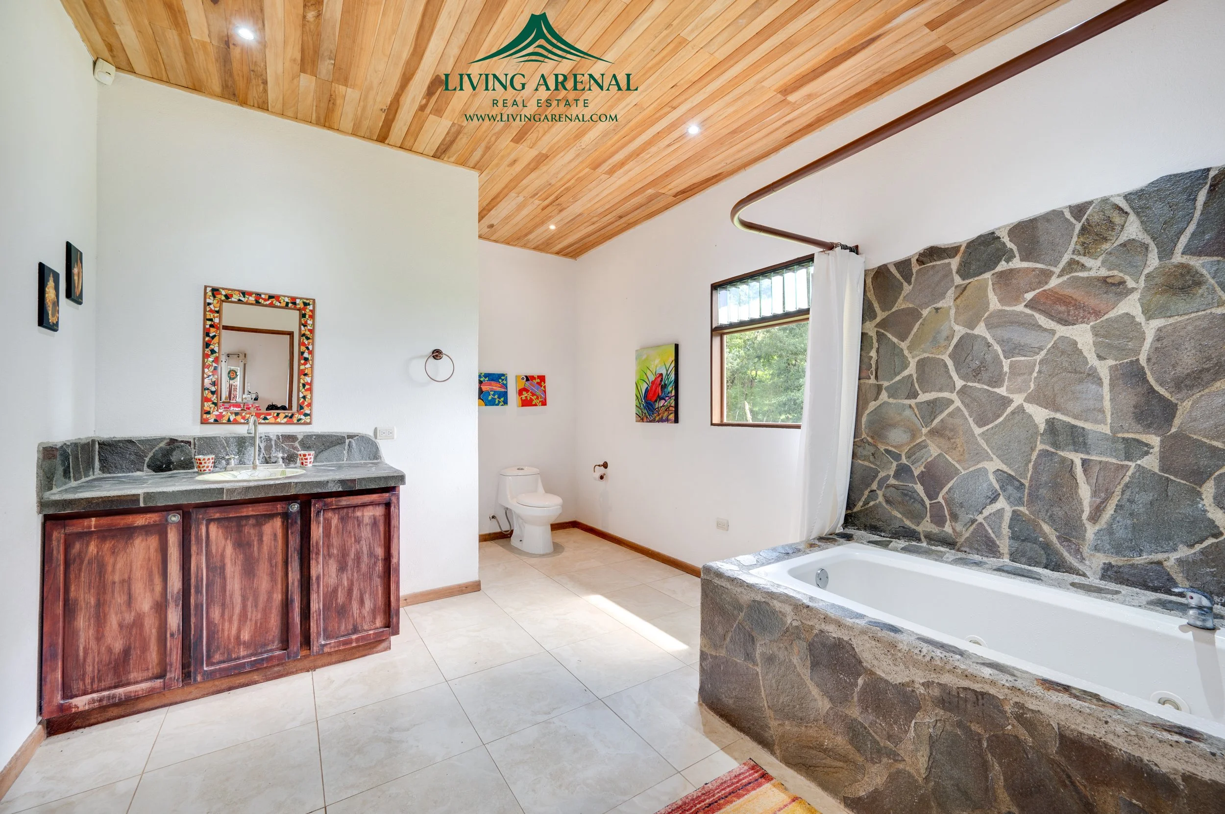 Bathroom with a stone bathtub, wooden vanity with a mirror, colorful wall art, window with greenery outside, white walls, wood ceiling, tiled floor.
