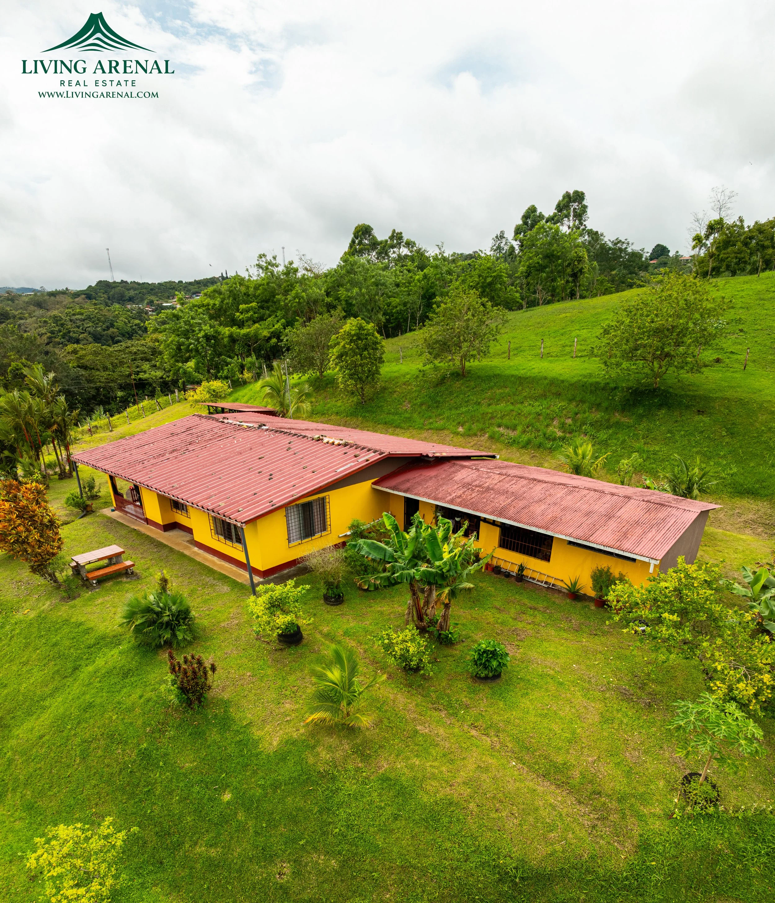 A yellow house with a red metal roof surrounded by lush green grass and trees in a rural area with hills in the background.