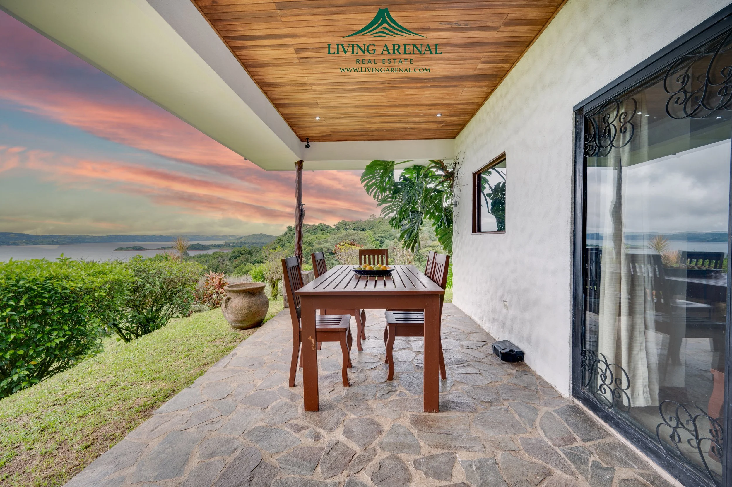 A covered patio overlooking a lake or river with lush green hills in the background. There is a wooden dining table with four chairs, a fruit bowl on the table, large potted plants nearby, and a stone floor. The patio ceiling is wooden with recessed