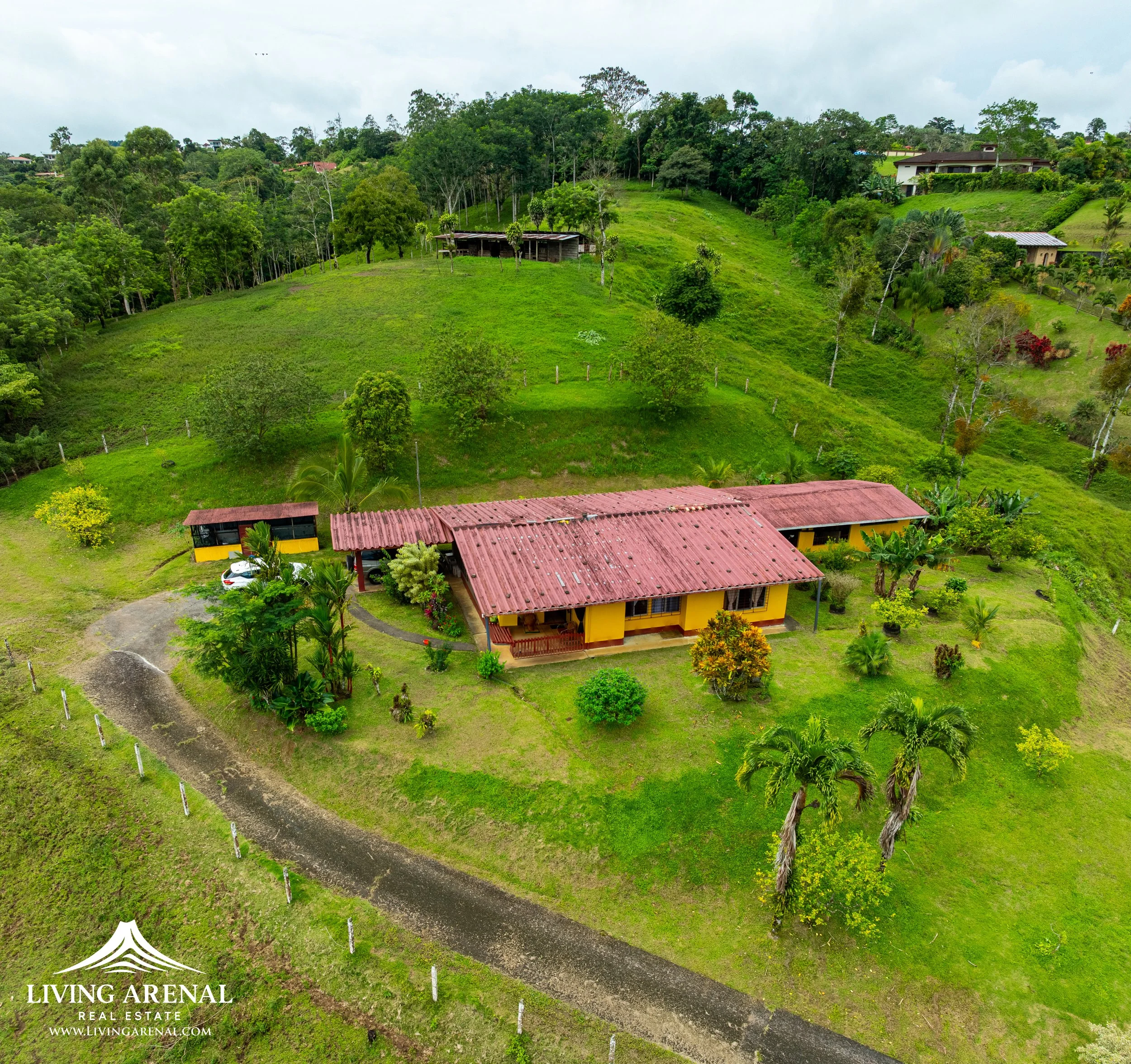 Aerial view of a yellow house with a red roof surrounded by green grass, trees, and hilly landscape. There is a driveway and several small buildings or structures nearby.