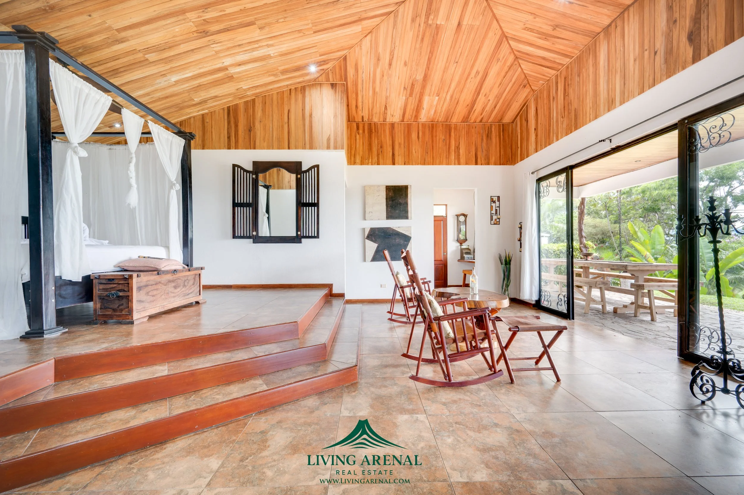 Interior of a spacious living area with wooden ceiling, white walls, and a view of a screened porch with outdoor seating, including rocking chairs and a picnic table, overlooking lush greenery.