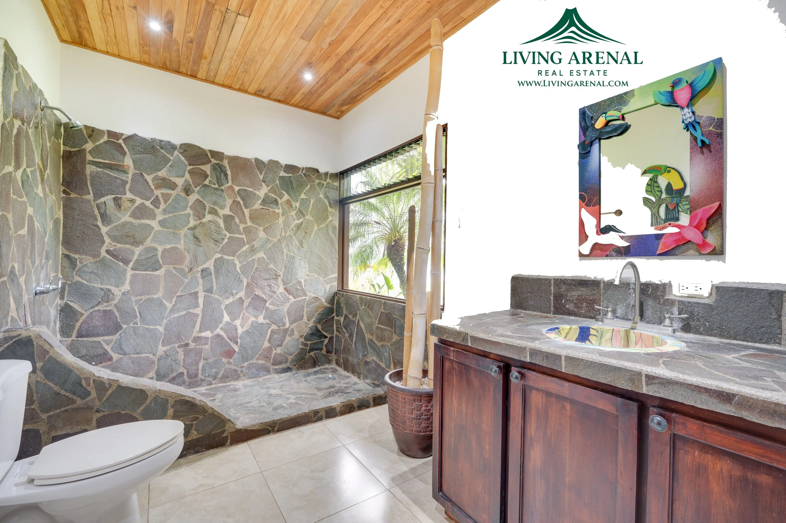 Bathroom with a stone shower area, wooden ceiling, window with view of greenery, wooden vanity with stone countertop, colorful mirror with 3 birds, potted plant, and toilet.