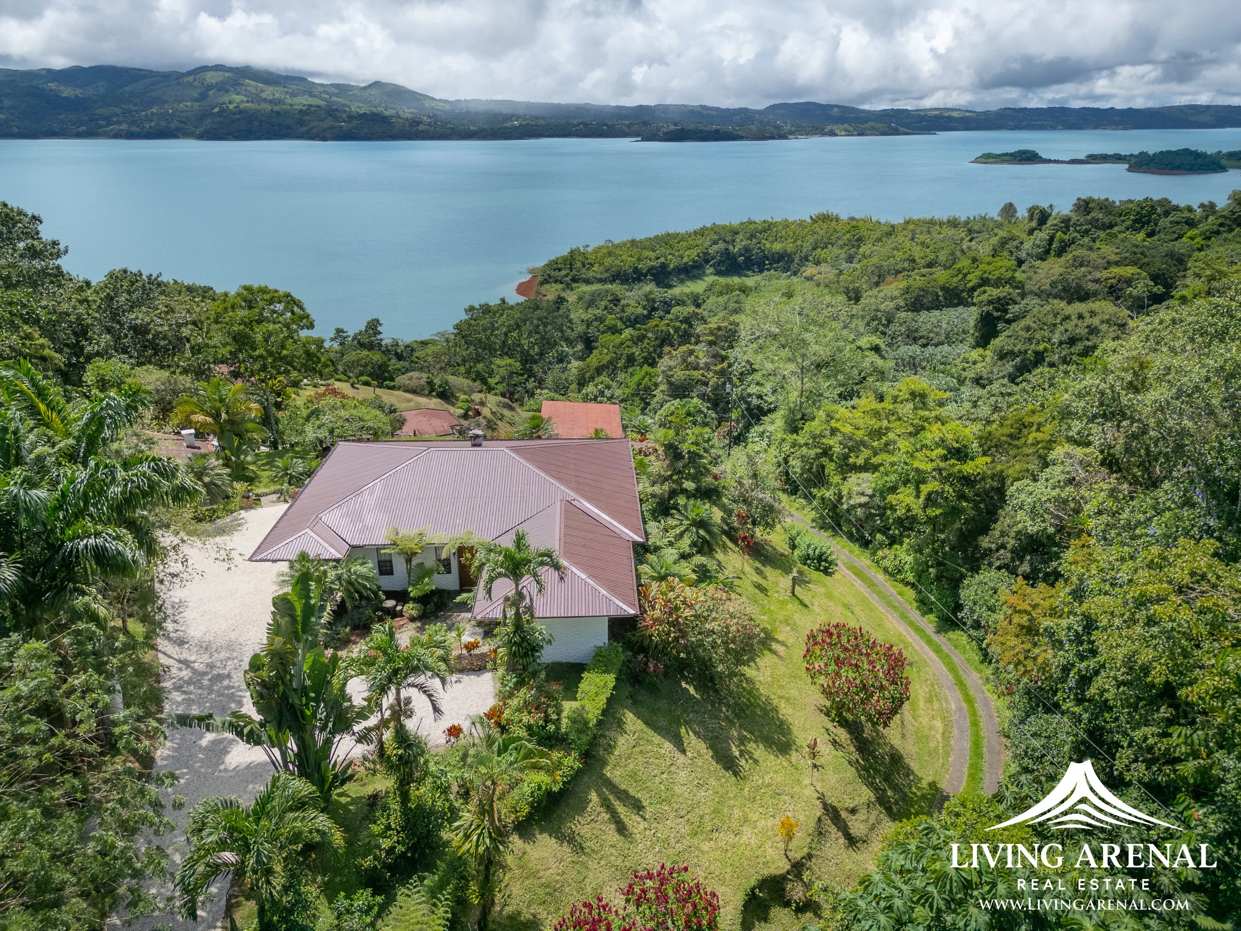 Aerial view of a house on a green landscape next to a large lake with mountains in the background.