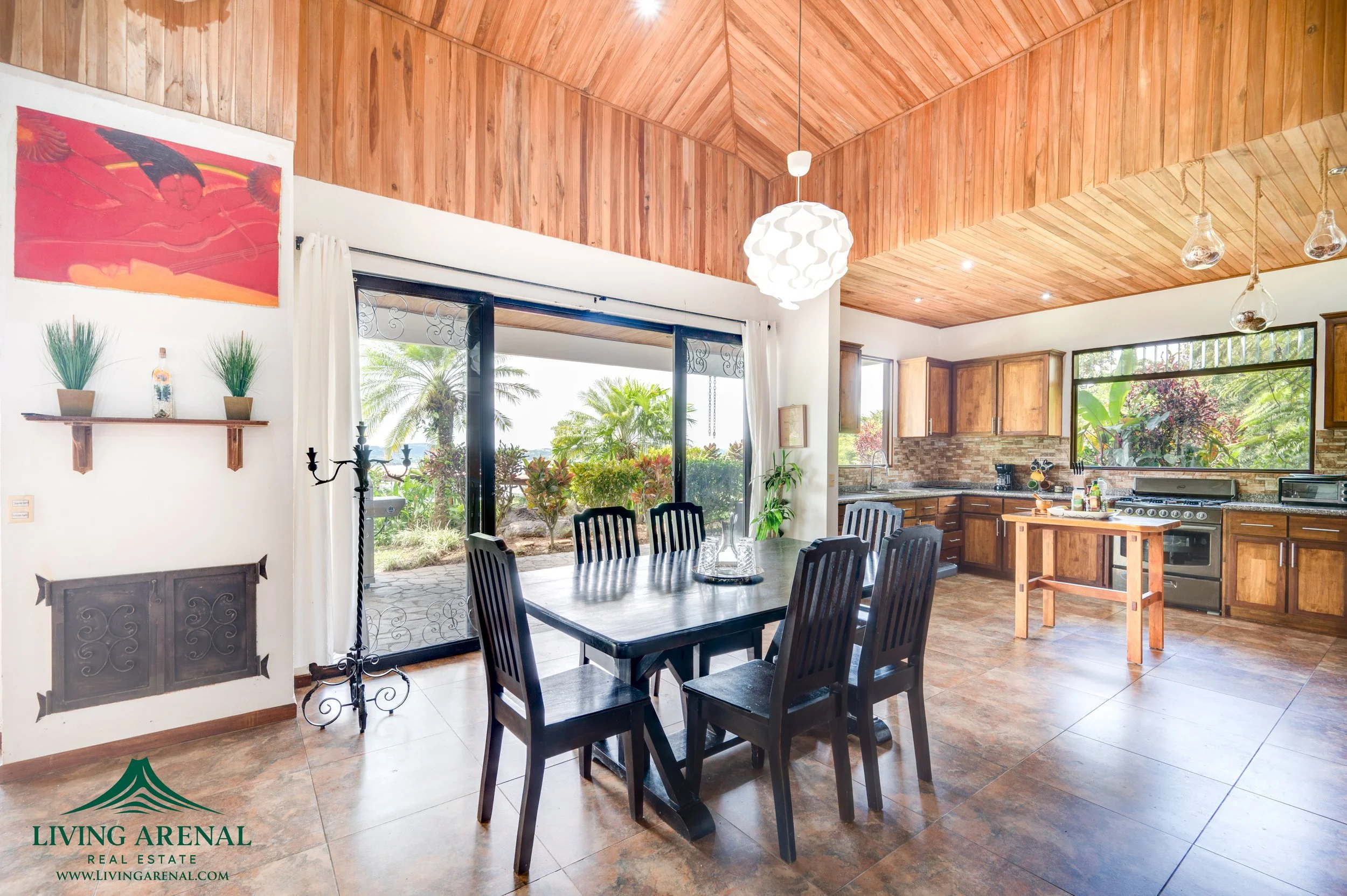 Open concept kitchen and dining area with wooden ceiling, black dining table with six chairs, large window, sliding glass door leading outside, and kitchen with wooden cabinets, stainless steel appliances, and island.
