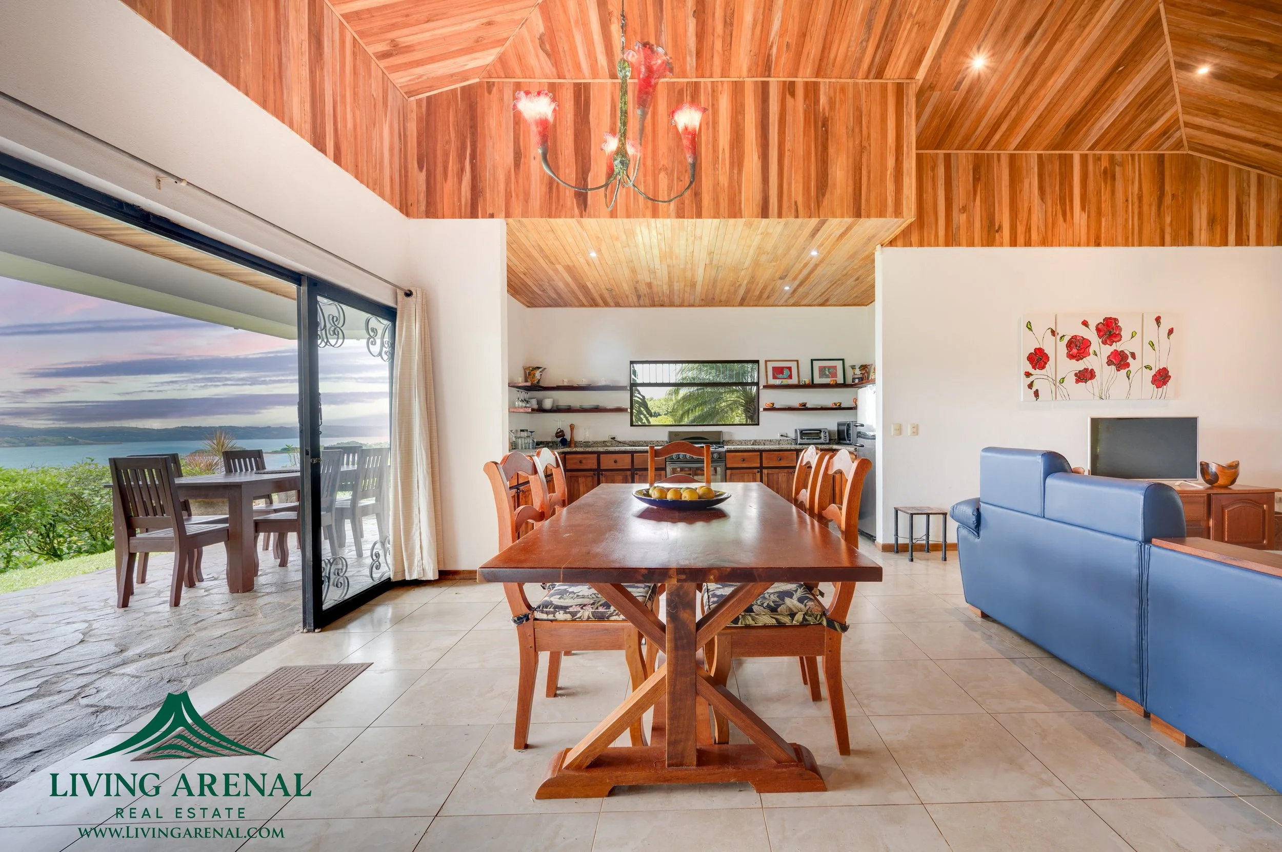 Interior view of a living and dining area with wooden vaulted ceiling, a dining table with eight chairs, a blue sofa, wall-mounted television, wall art, and sliding glass door leading to an outdoor patio with a view of greenery and water.